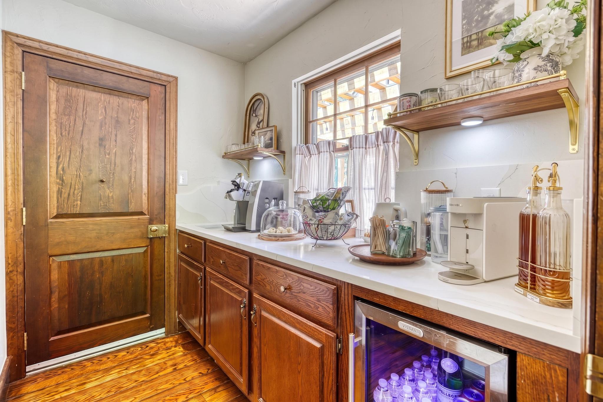 Pantry coffee and tea station with marble counter, brass-lit floating shelves, and stocked mini-fridge