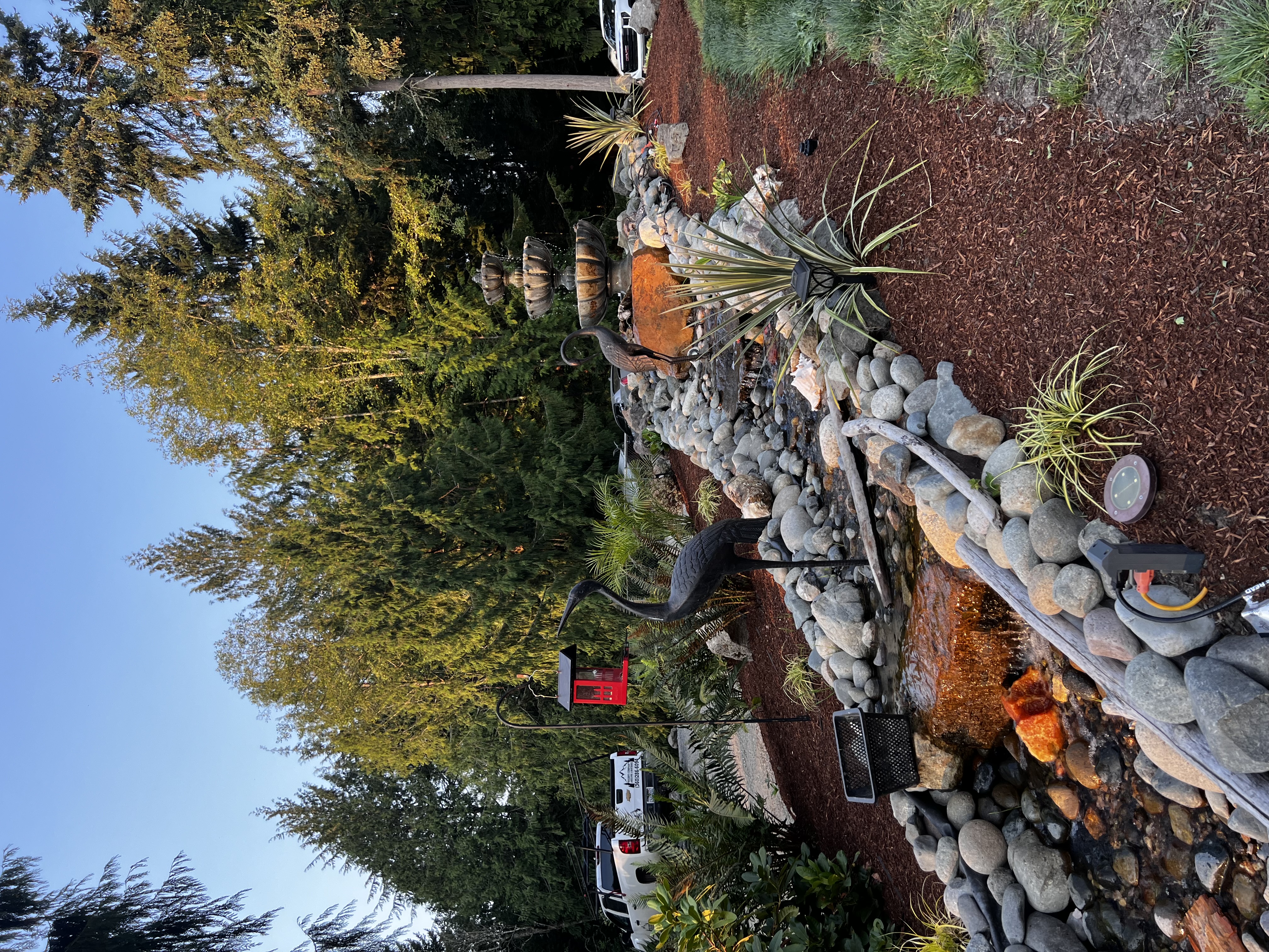 Stone dry creek bed water feature with river rocks, mulch, and a crane sculpture backed by evergreens