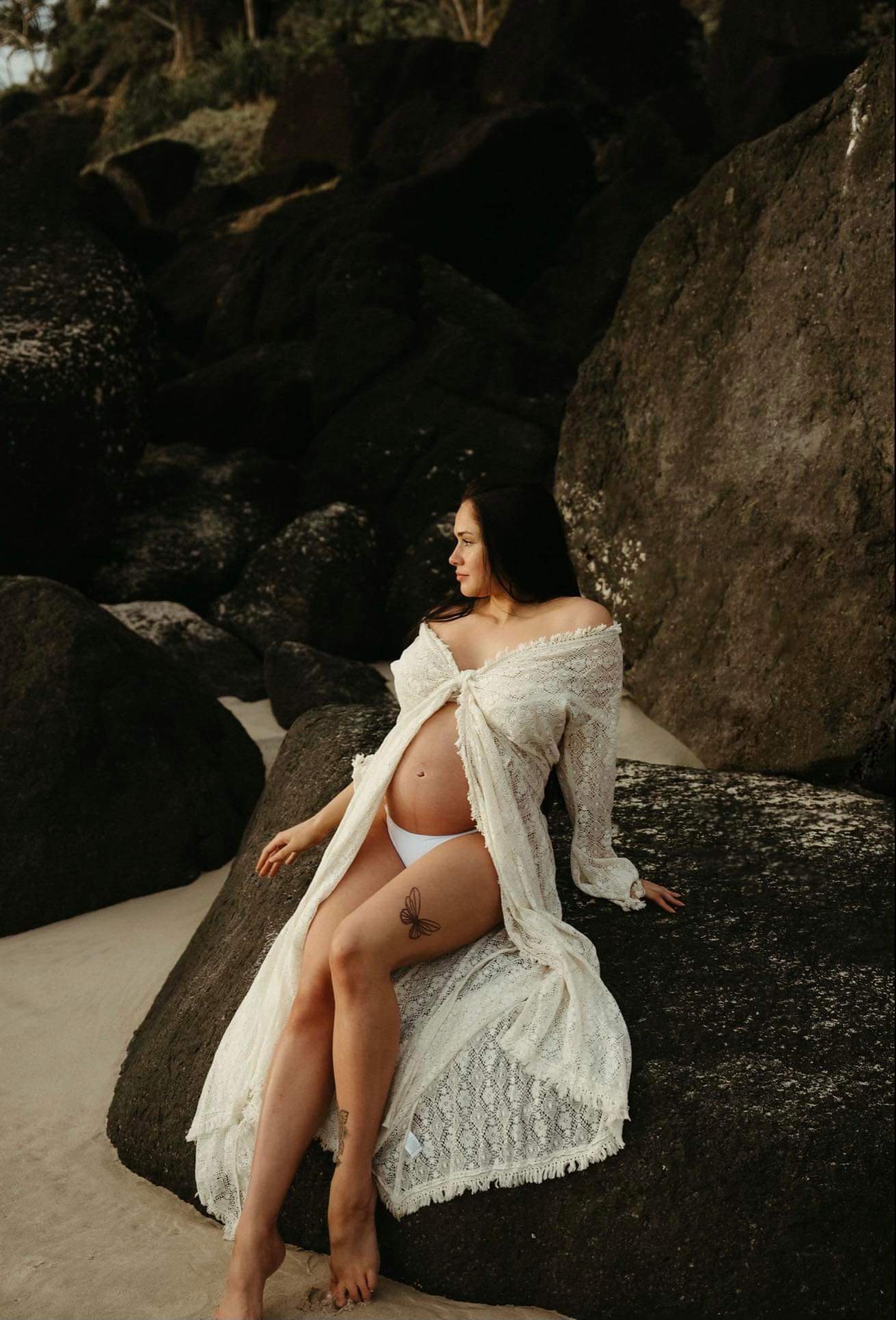 A pregnant woman in a white lace robe resting her hand on her baby bump while sitting on dark coastal rocks at the beach
