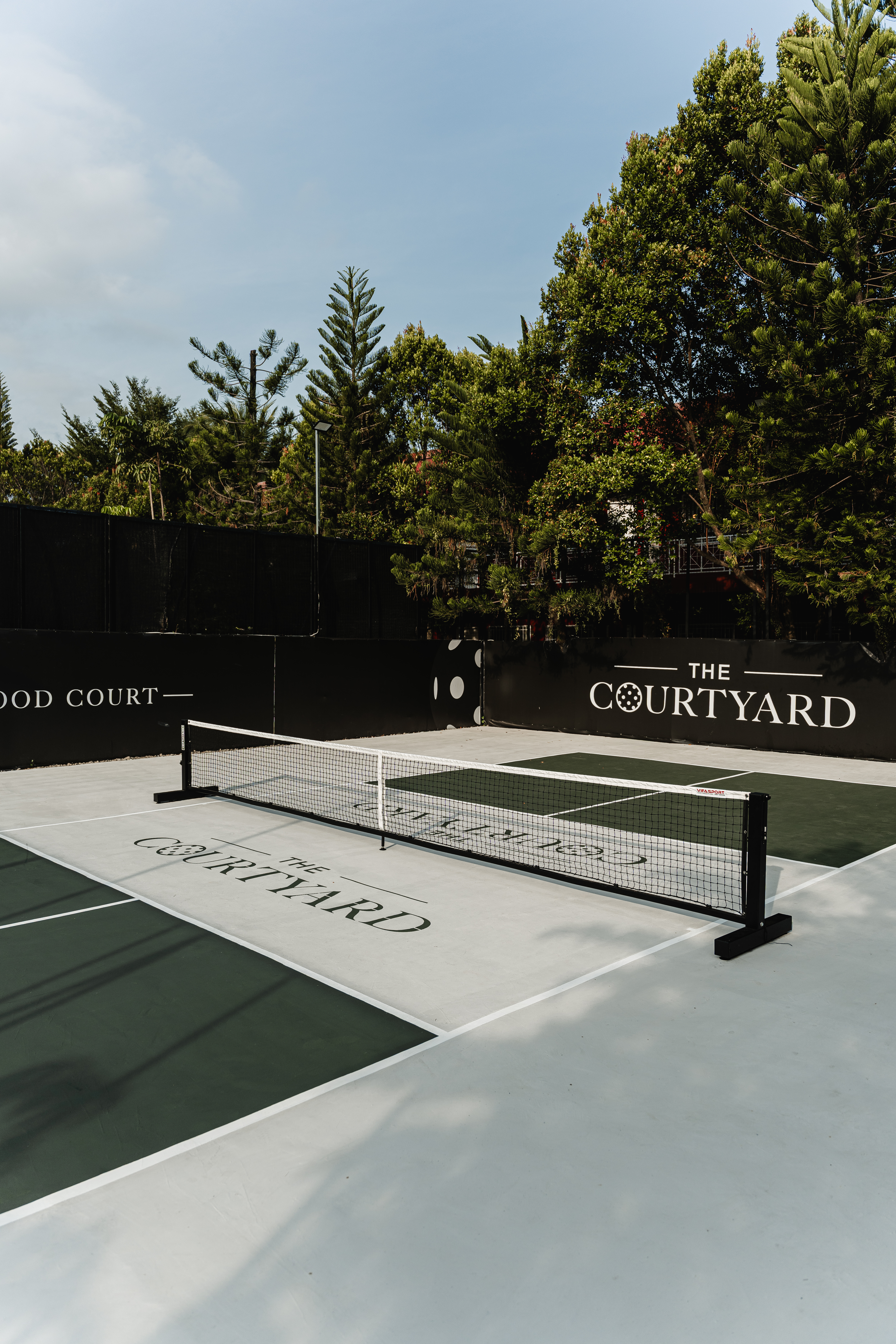 The Courtyard outdoor pickleball court in daylight, with the branded perimeter wall and trees behind