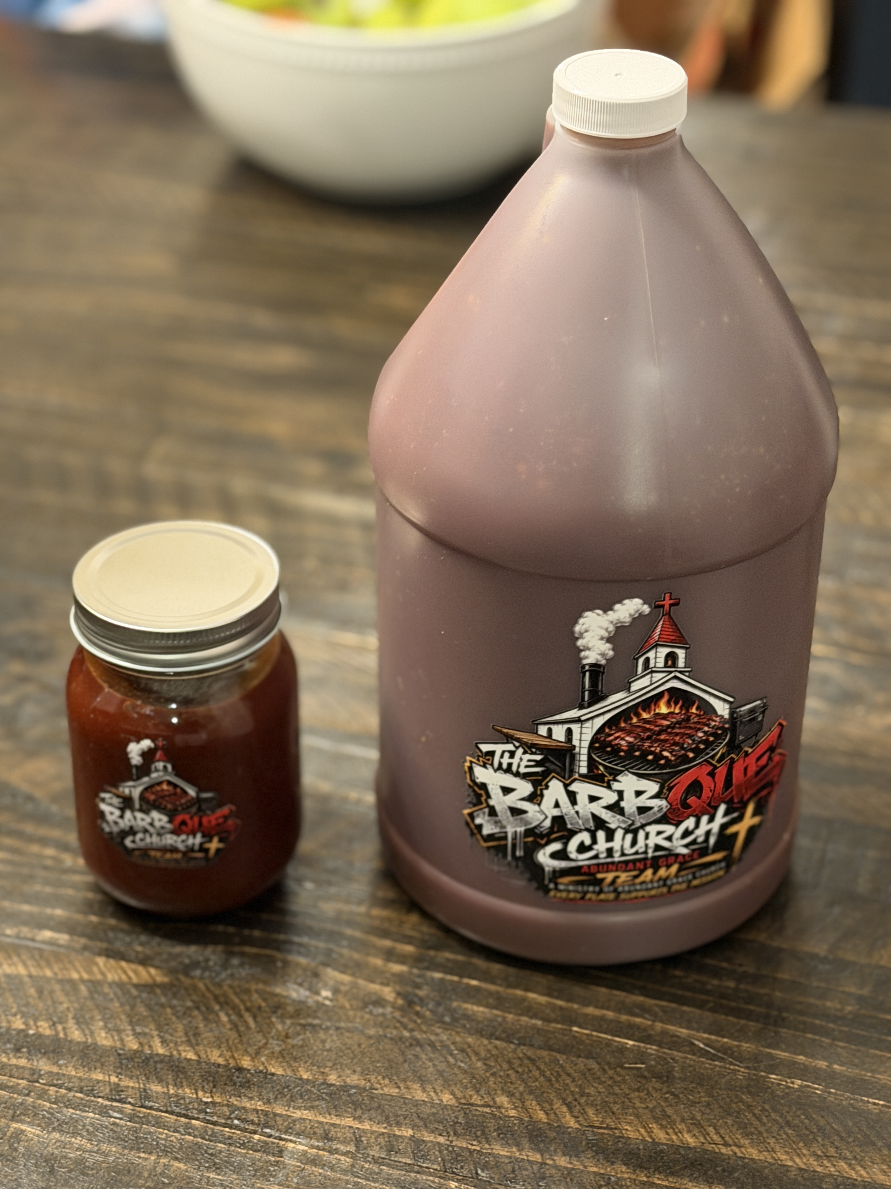 The Barbeque Church team barbecue sauce — a mason jar and gallon jug labeled "Every plate supports the mission"