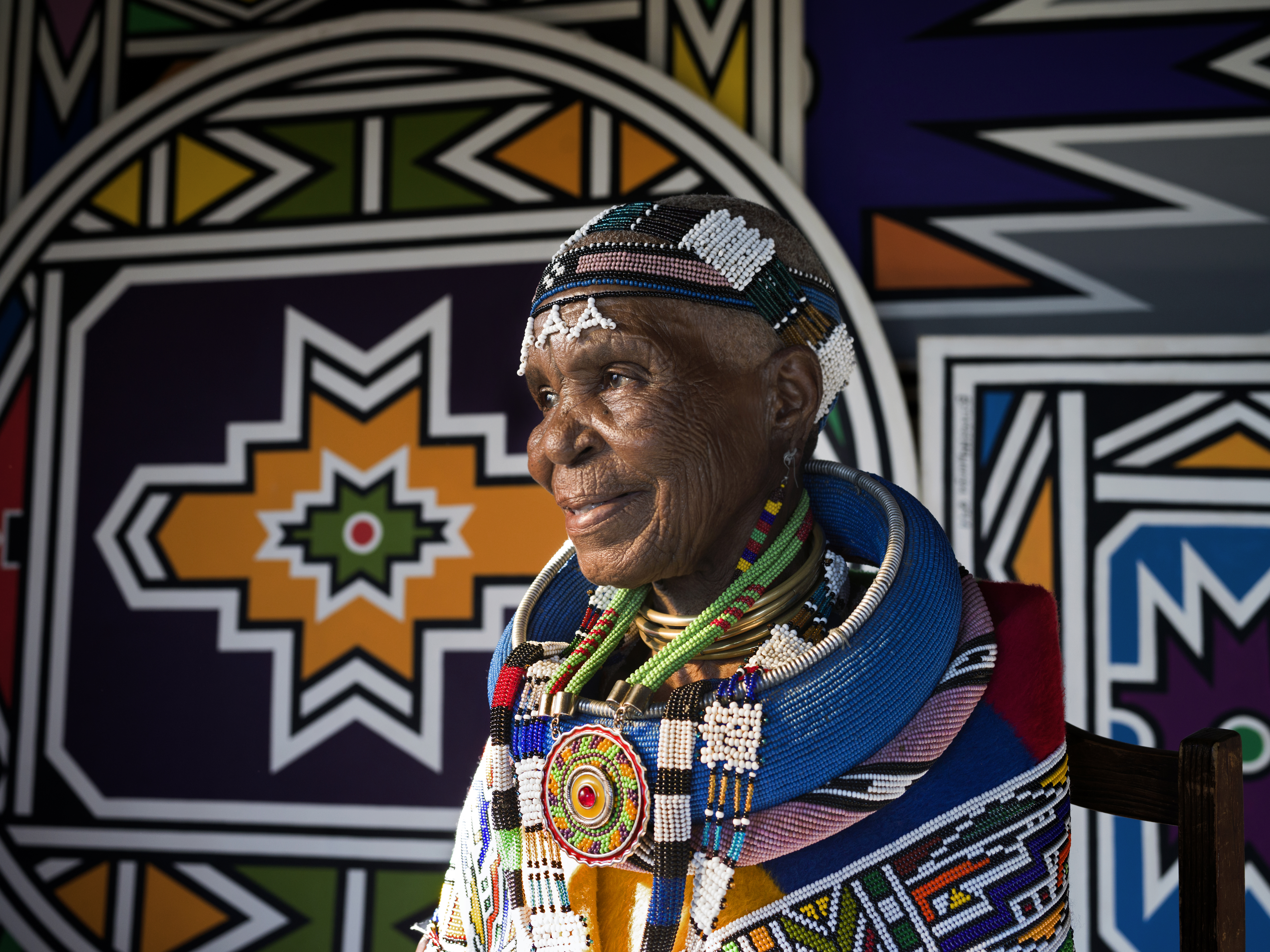 Esther Mahlangu wearing traditional Ndebele beadwork and neck rings, positioned against a background of her signature geometric mural art.