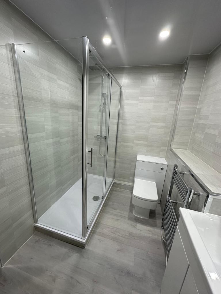 A modern renovated bathroom in Edinburgh with a walk-in shower, white toilet, chrome heated towel rail, vanity unit and grey wood-effect flooring, fitted by Calico Joinery