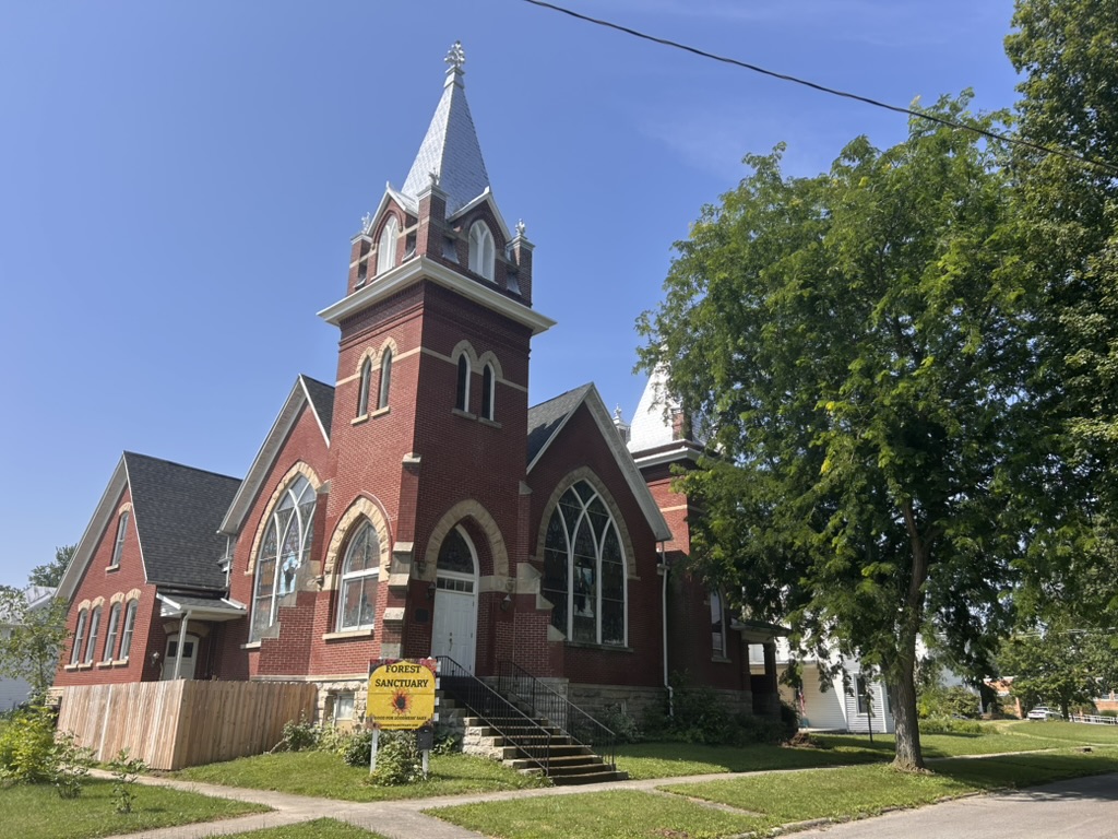 Forest Sanctuary — the red-brick chapel in Forest, Ohio, with its yard sign reading Good for Someone Else