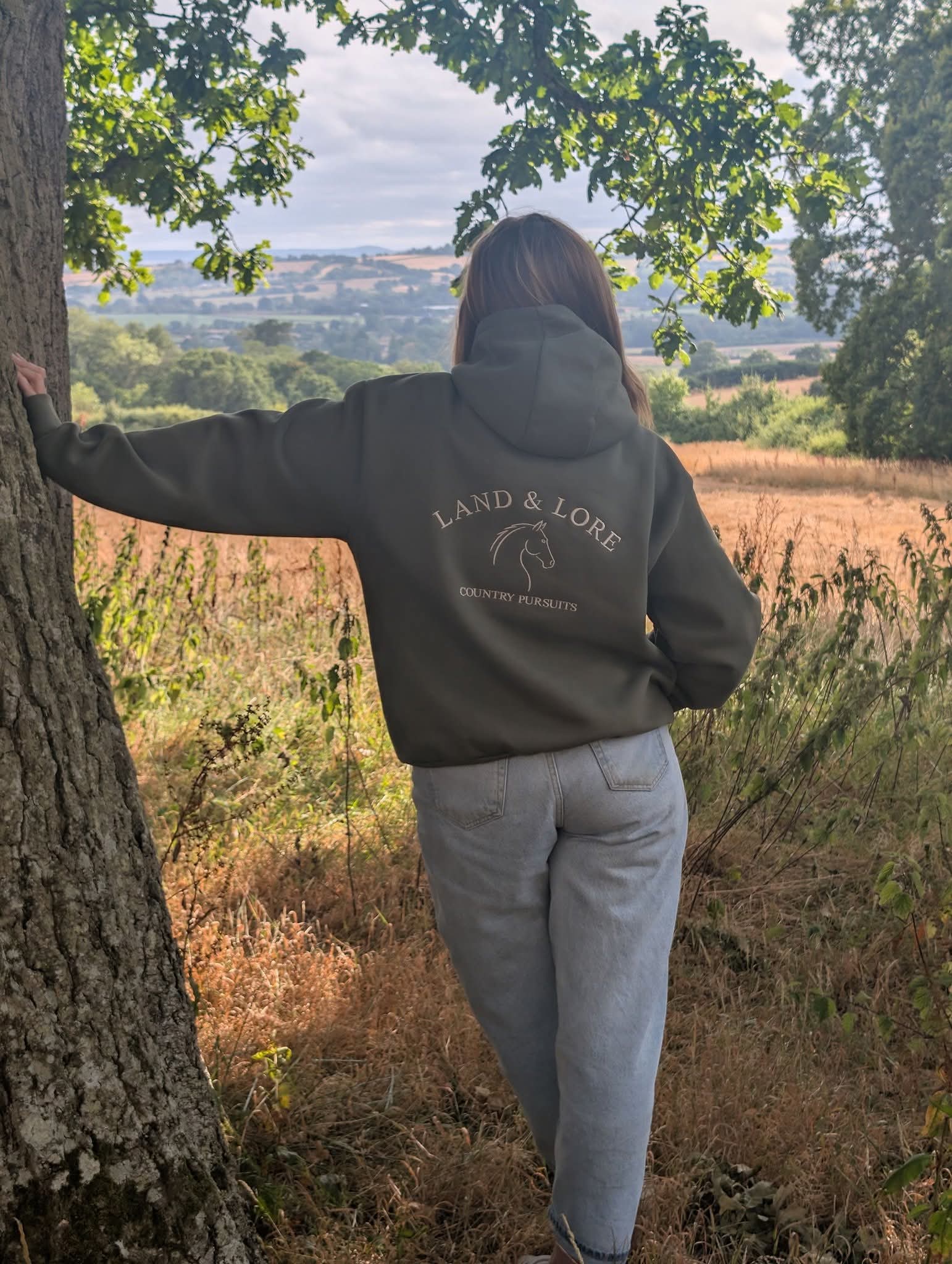 Person in olive Land & Lore hoodie leaning against a tree