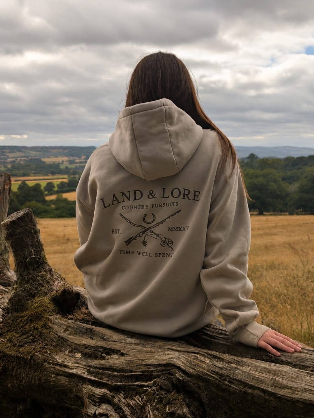 Person in sand Land & Lore hoodie sitting on a tree trunk in an open field