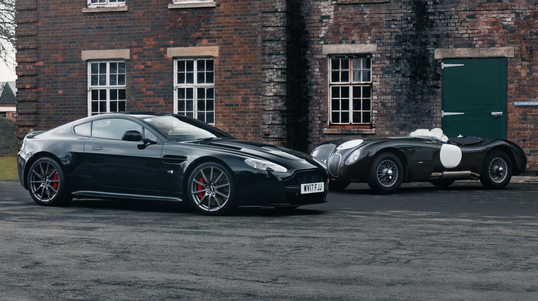 A modern Aston Martin and a classic racing car parked outside a red-brick building