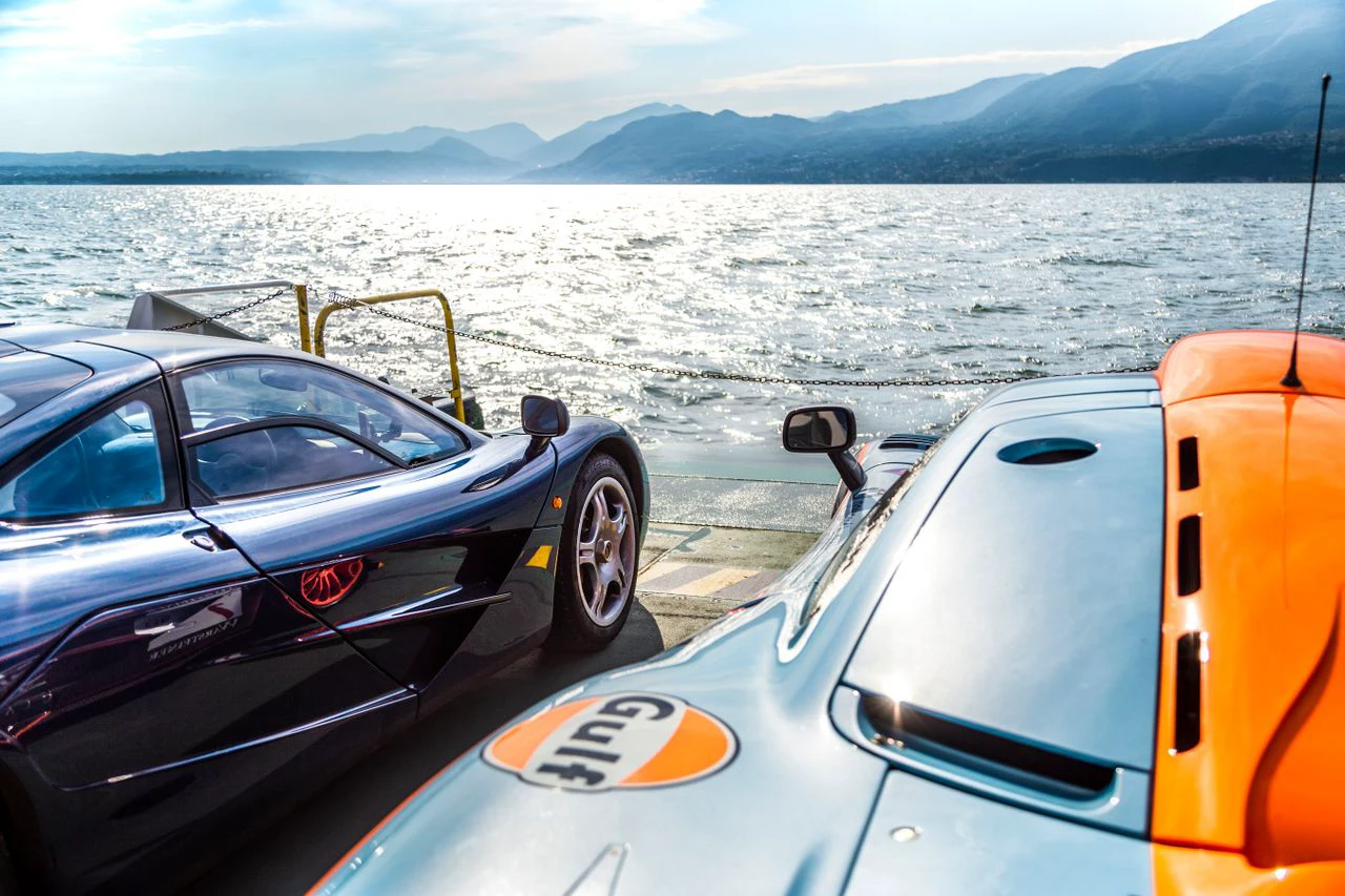 Two McLaren F1s on a ferry deck against a lake and mountains