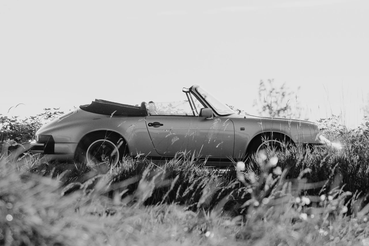 A vintage Porsche 911 Cabriolet parked in tall grass, backlit in black and white