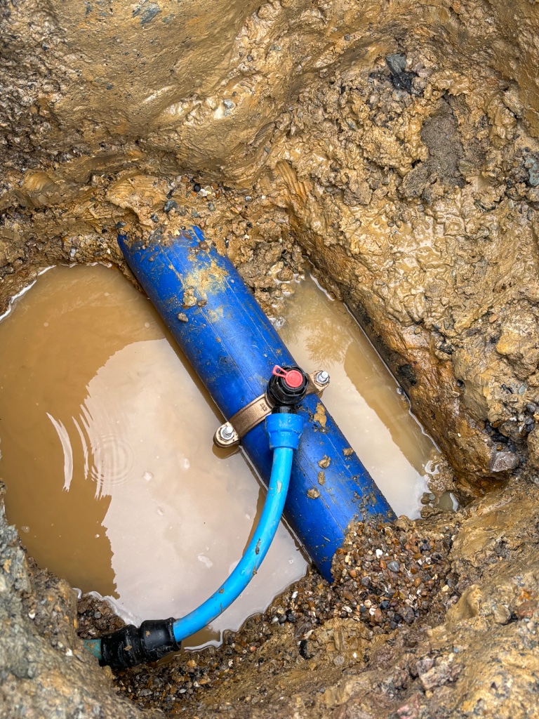 Underground blue water pipe repair with brass saddle clamp in excavated trench by Subsoil Land & Water in Battle East Sussex