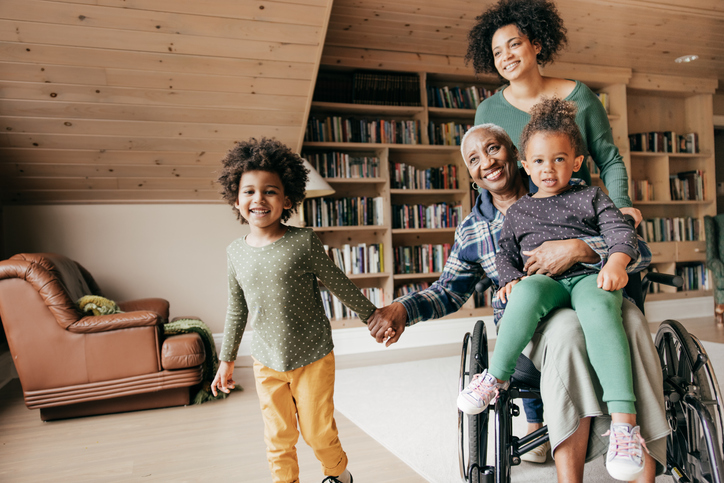 A multi-generational family gathered in a light-filled room — a grandmother in a wheelchair holding a child, a younger woman resting a hand on her shoulder