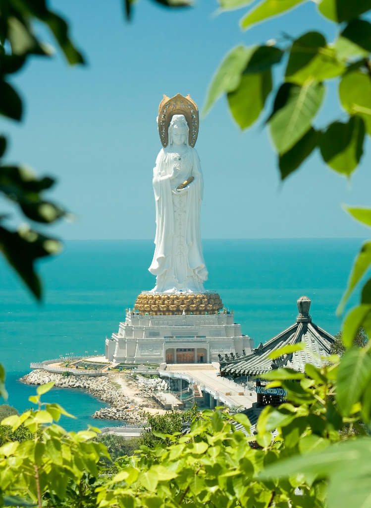 A white statue of Guanyin, the Bodhisattva of Compassion, rising above a calm turquoise sea