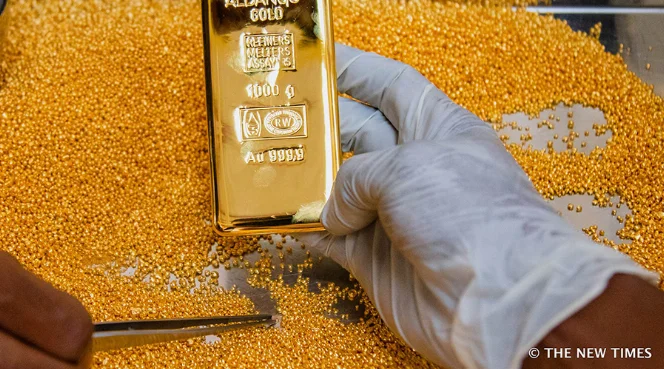 A gloved hand holding a polished gold bullion bar over gold granules