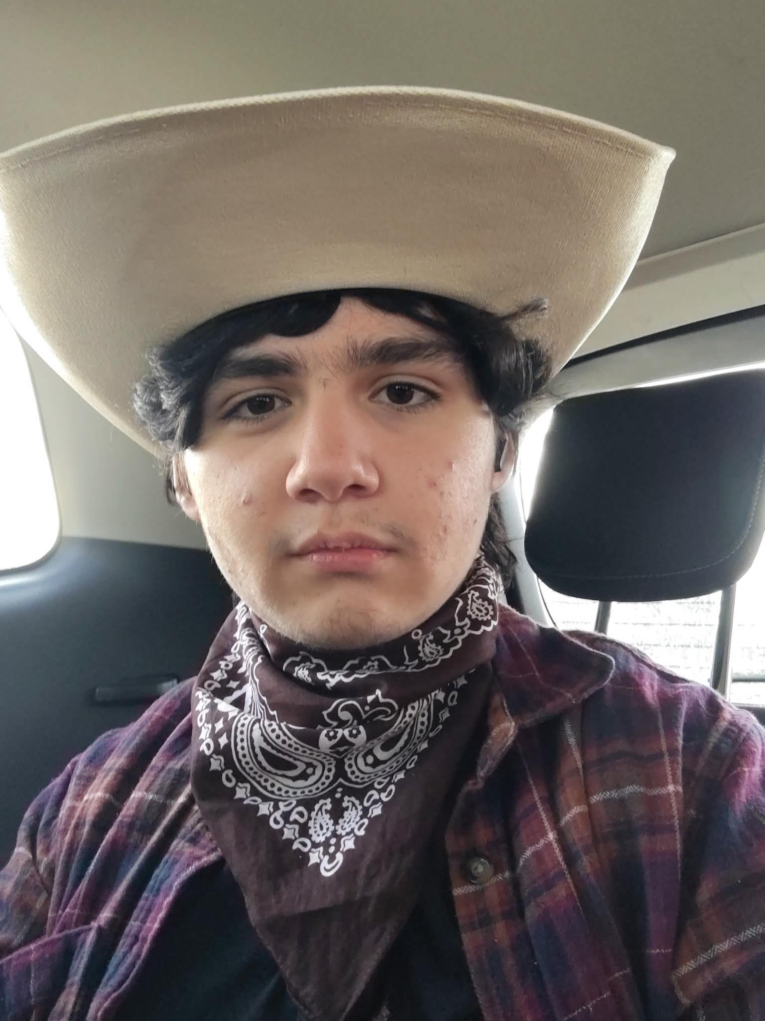 Portrait of Doni E. wearing a tan cowboy hat and paisley bandana inside a vehicle, soft natural lighting