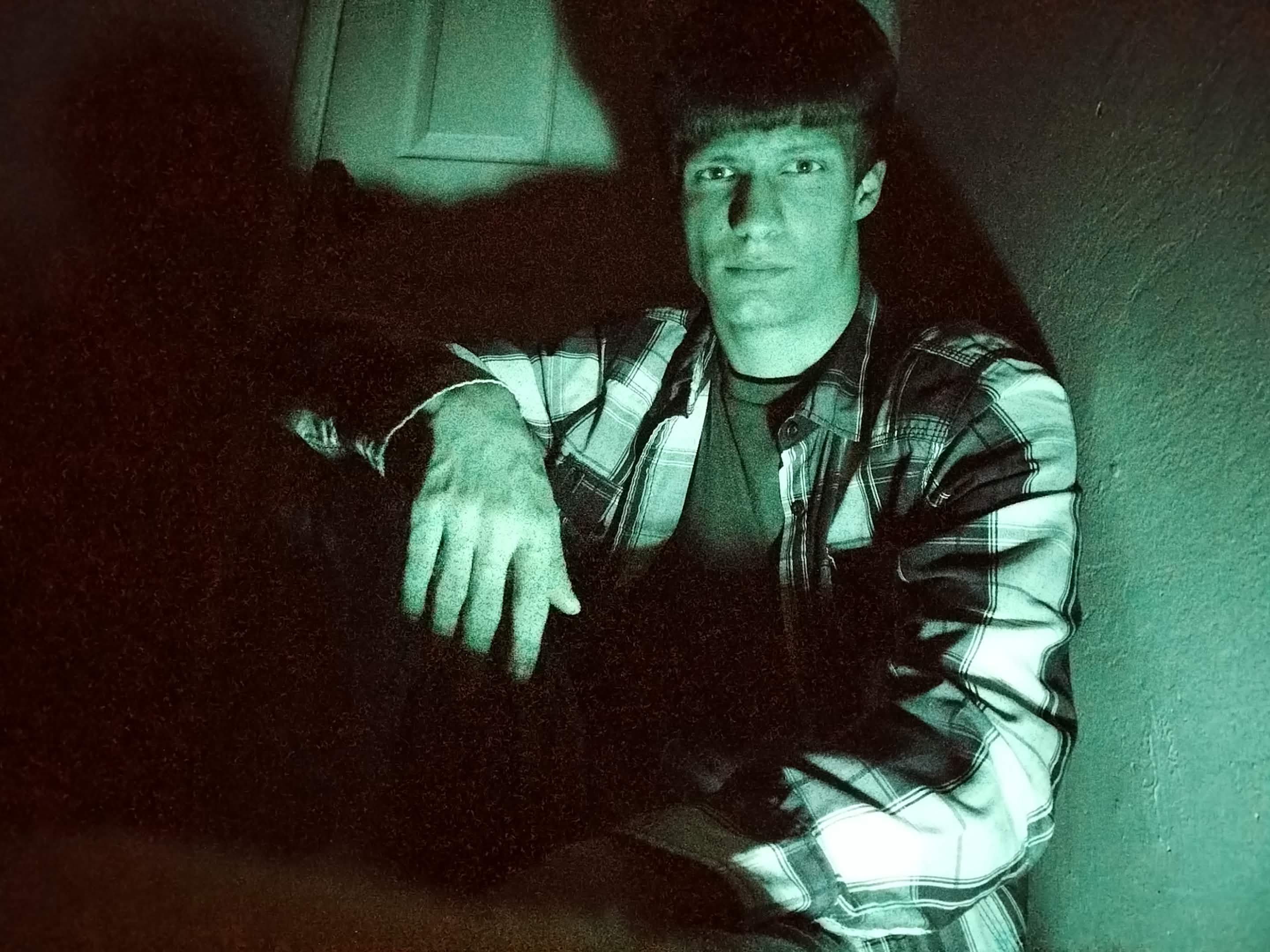 Portrait of Nathan Jameson under vibrant green lighting, wearing an unbuttoned plaid flannel shirt over a dark t-shirt