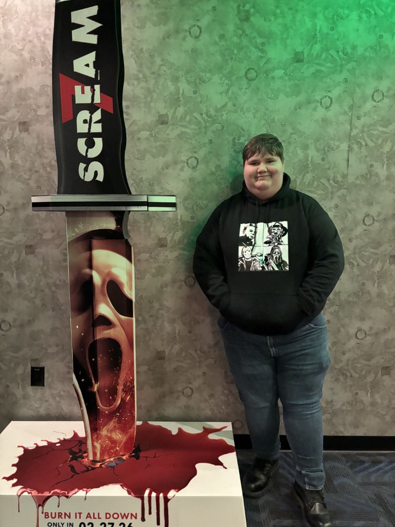 Portrait of Nik Lynch standing next to a Scream movie promotional display, wearing a black horror icon hoodie and jeans