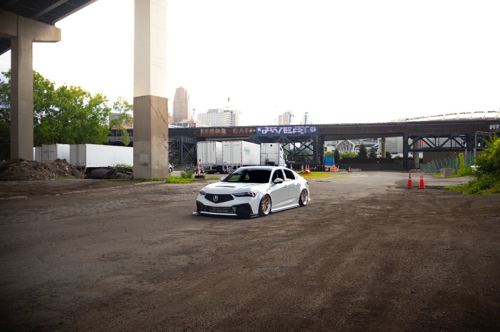 Lowered white Acura sedan parked under a concrete overpass with graffiti