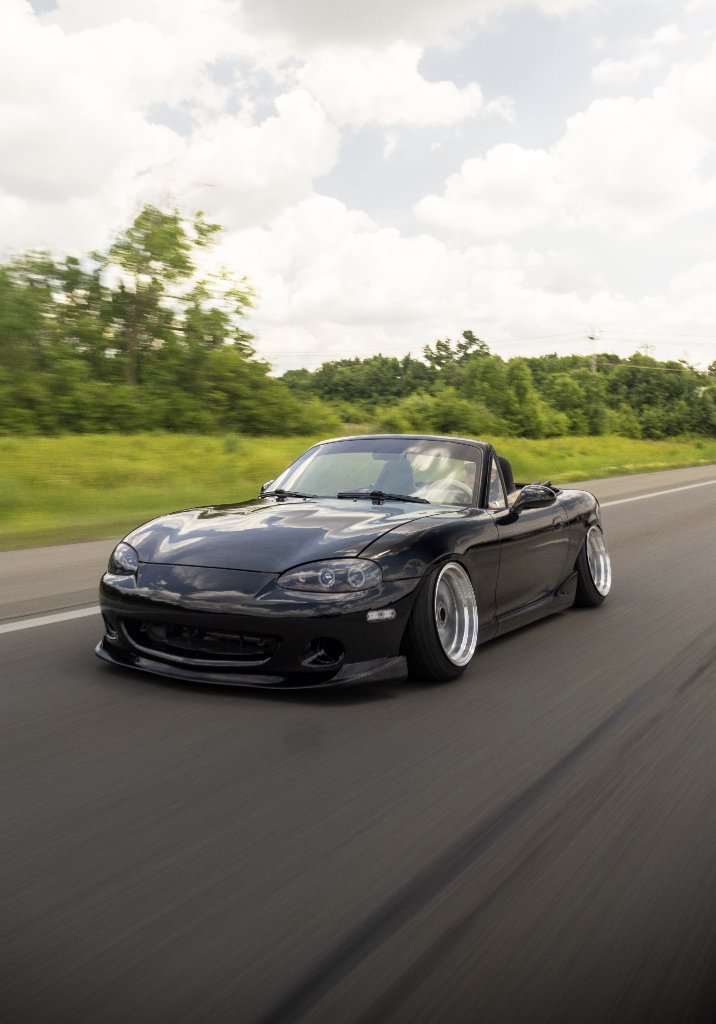Black Mazda Miata convertible motion blur rolling shot on road with trees