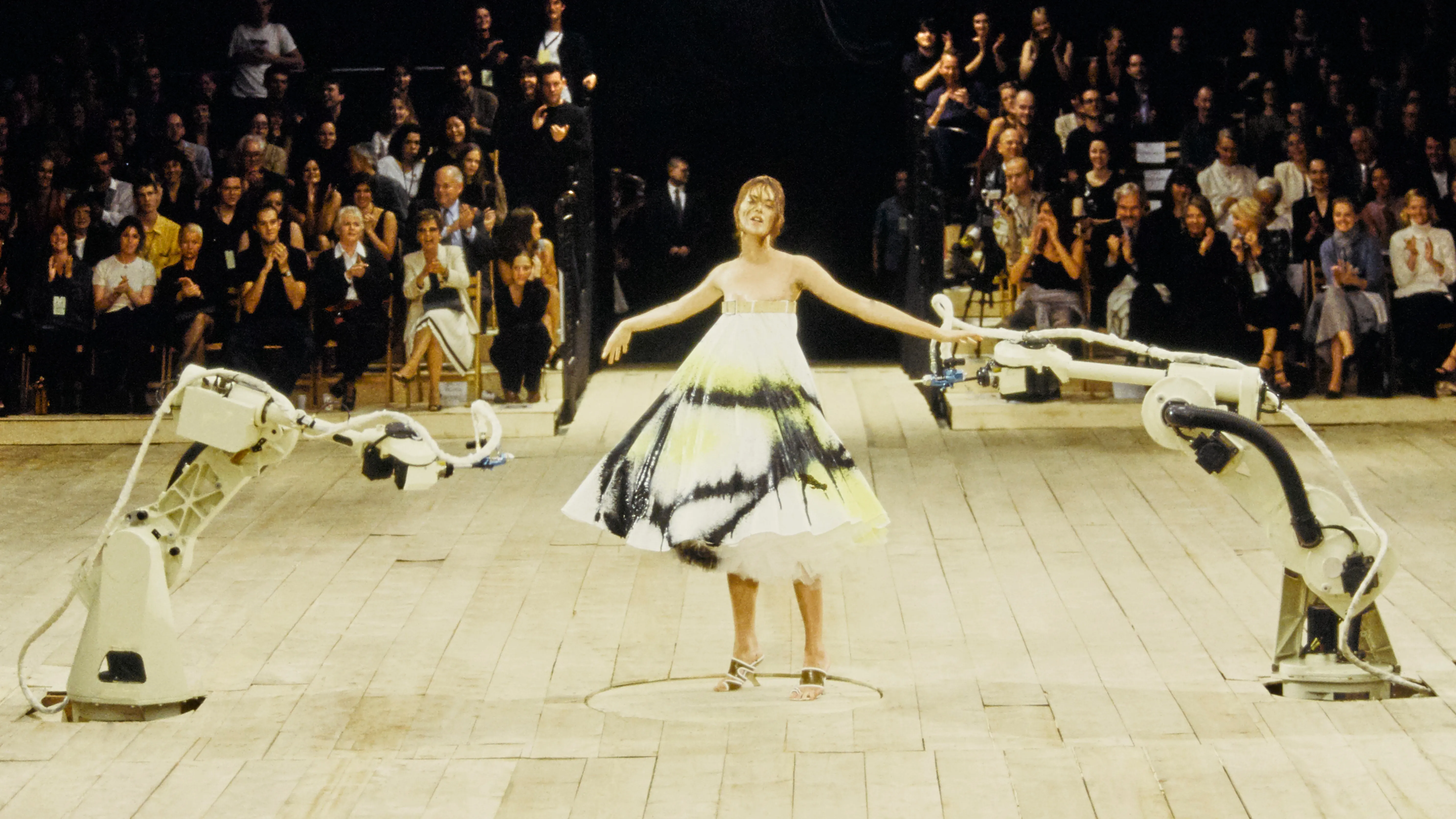 Shalom Harlow stands between two industrial robotic arms on a wooden runway as they spray paint across her white dress