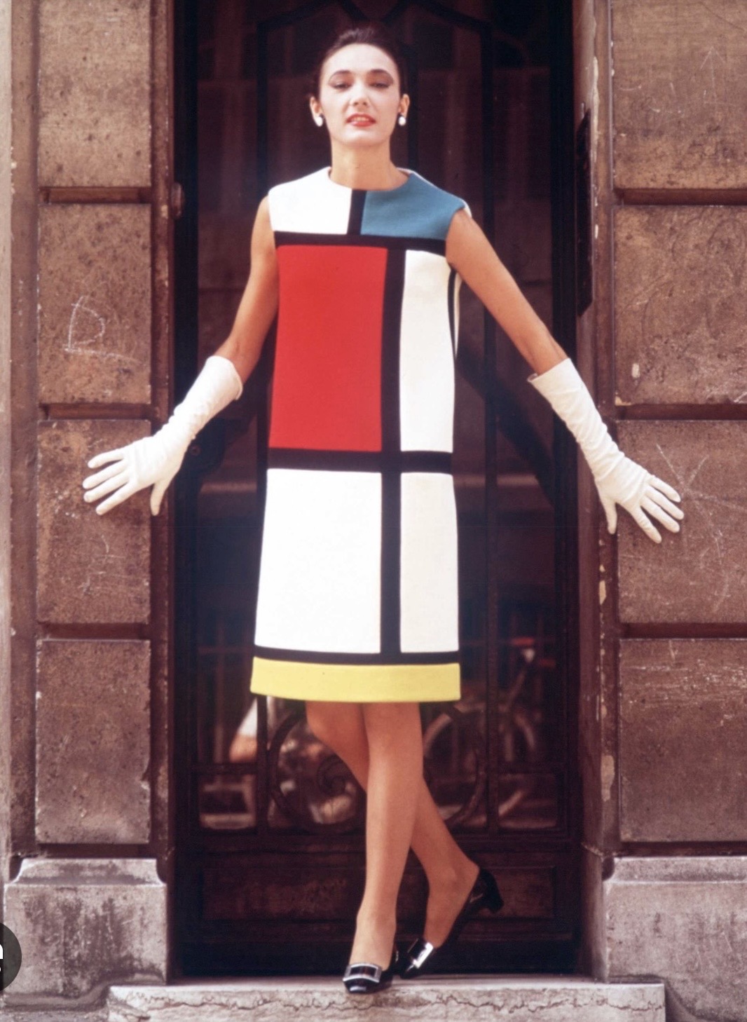 Yves Saint Laurent's 1965 Mondrian dress, a sleeveless shift dress with bold black lines dividing blocks of white, red, blue, and yellow, photographed in front of a wooden doorway.