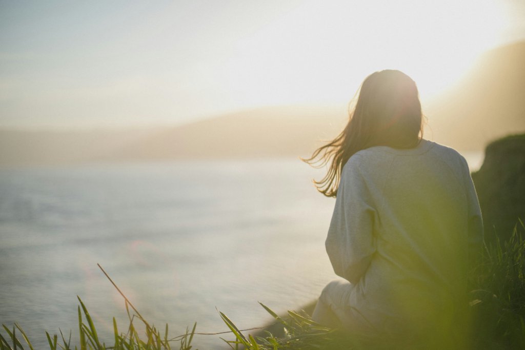 Person sitting on a grassy cliff overlooking the ocean at sunset