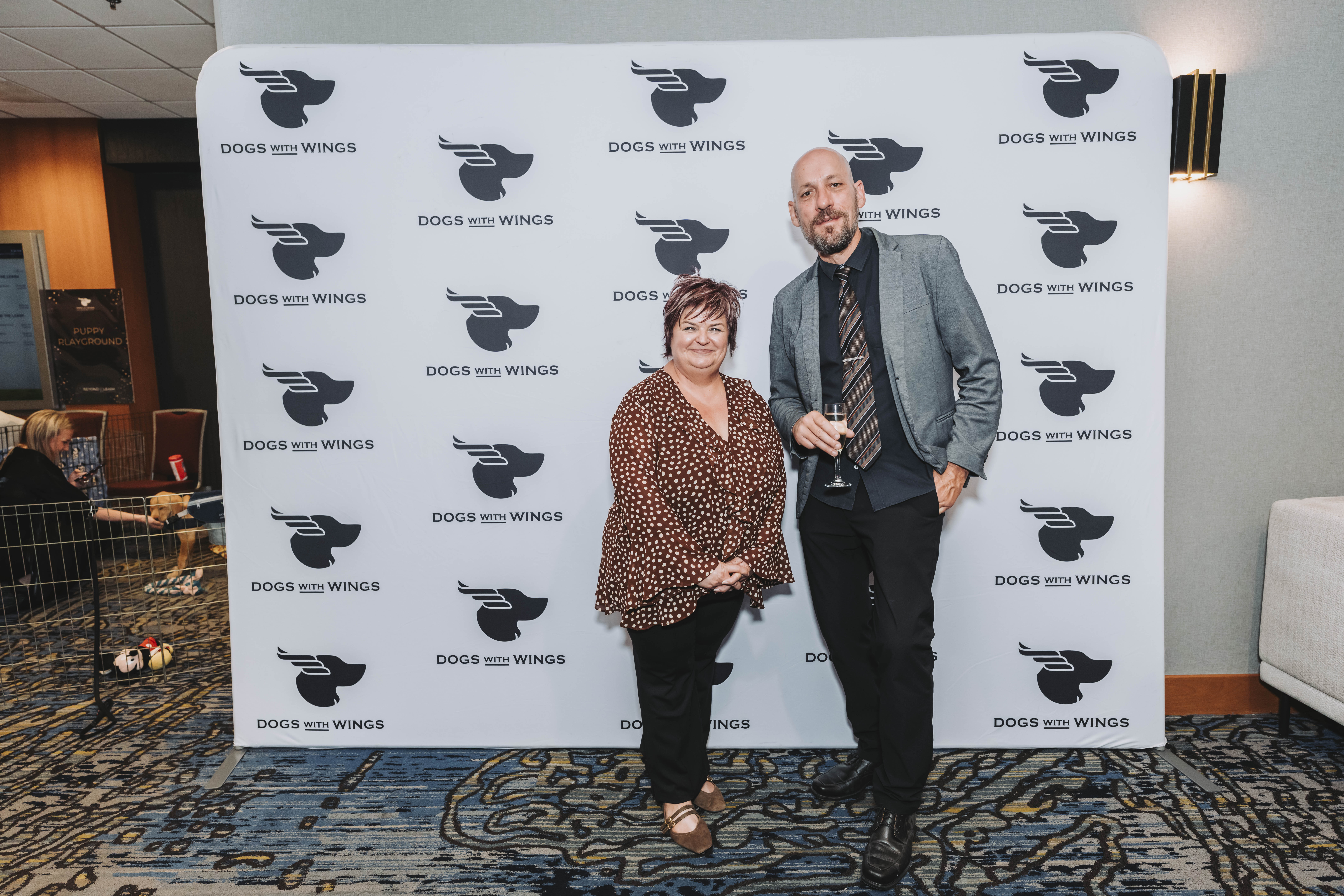 A couple posing at the Dogs With Wings backdrop by the puppy pen