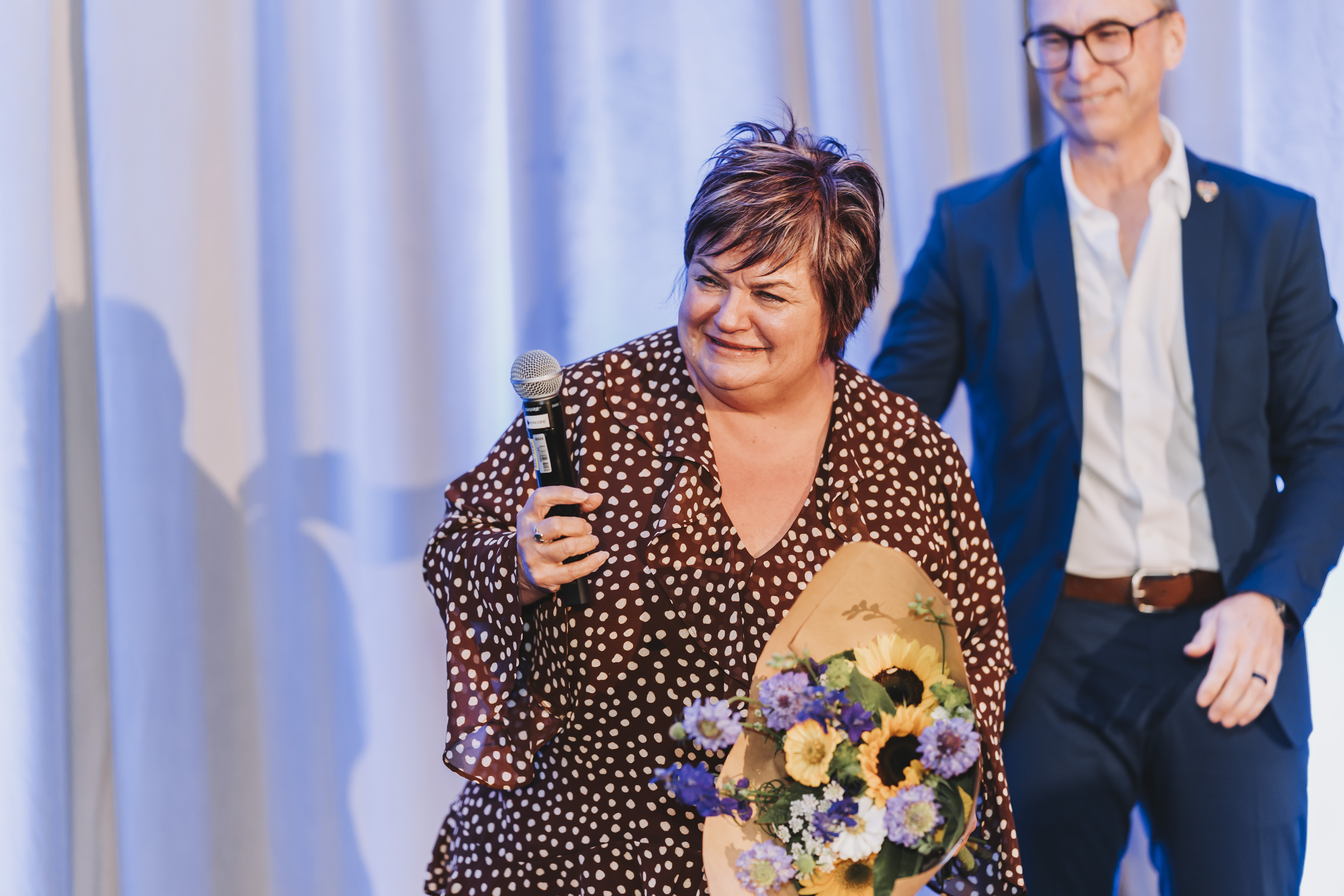 A guest on stage holding a microphone and a bouquet of sunflowers