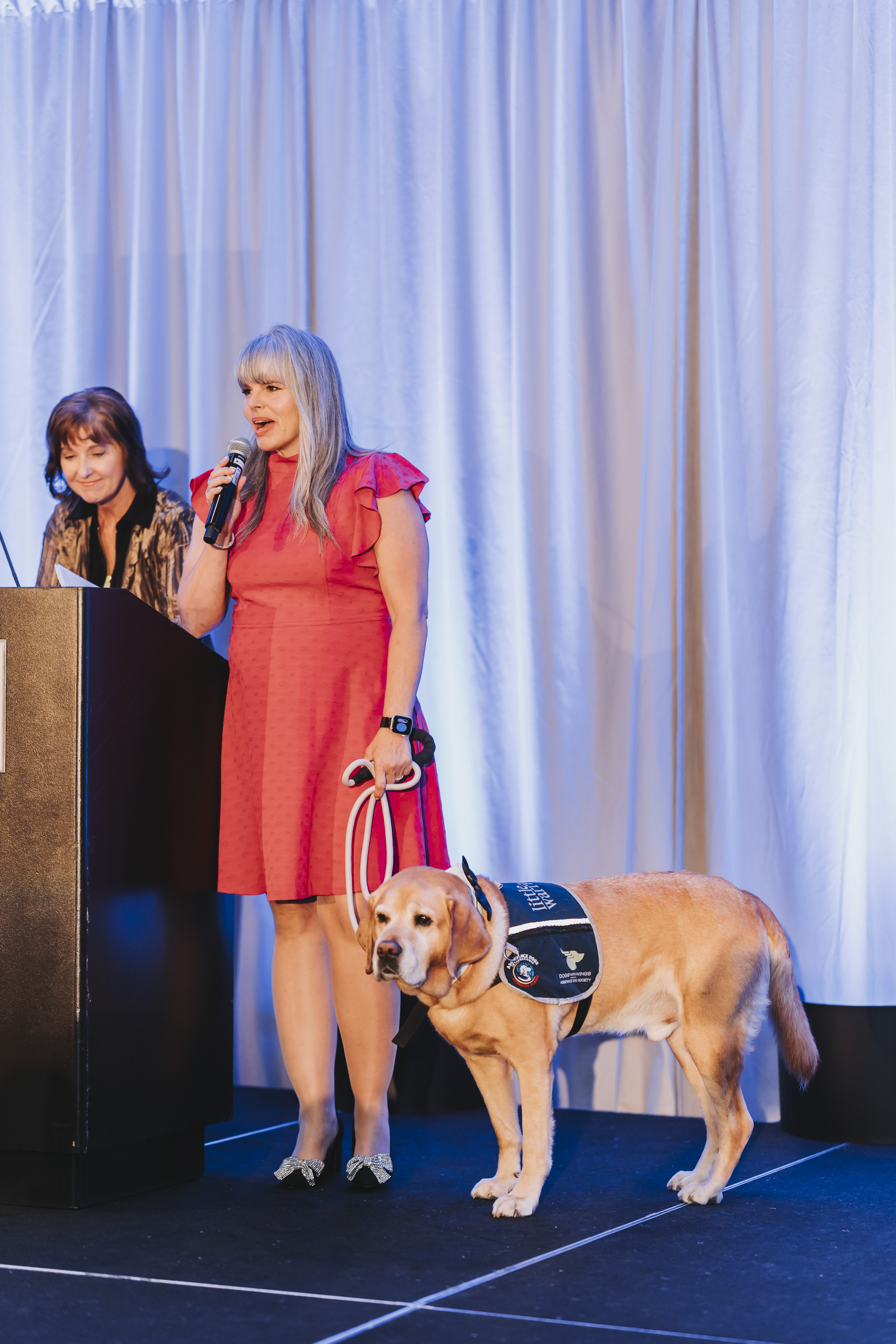 A guest speaking on stage with a yellow Labrador in a Little Wings vest