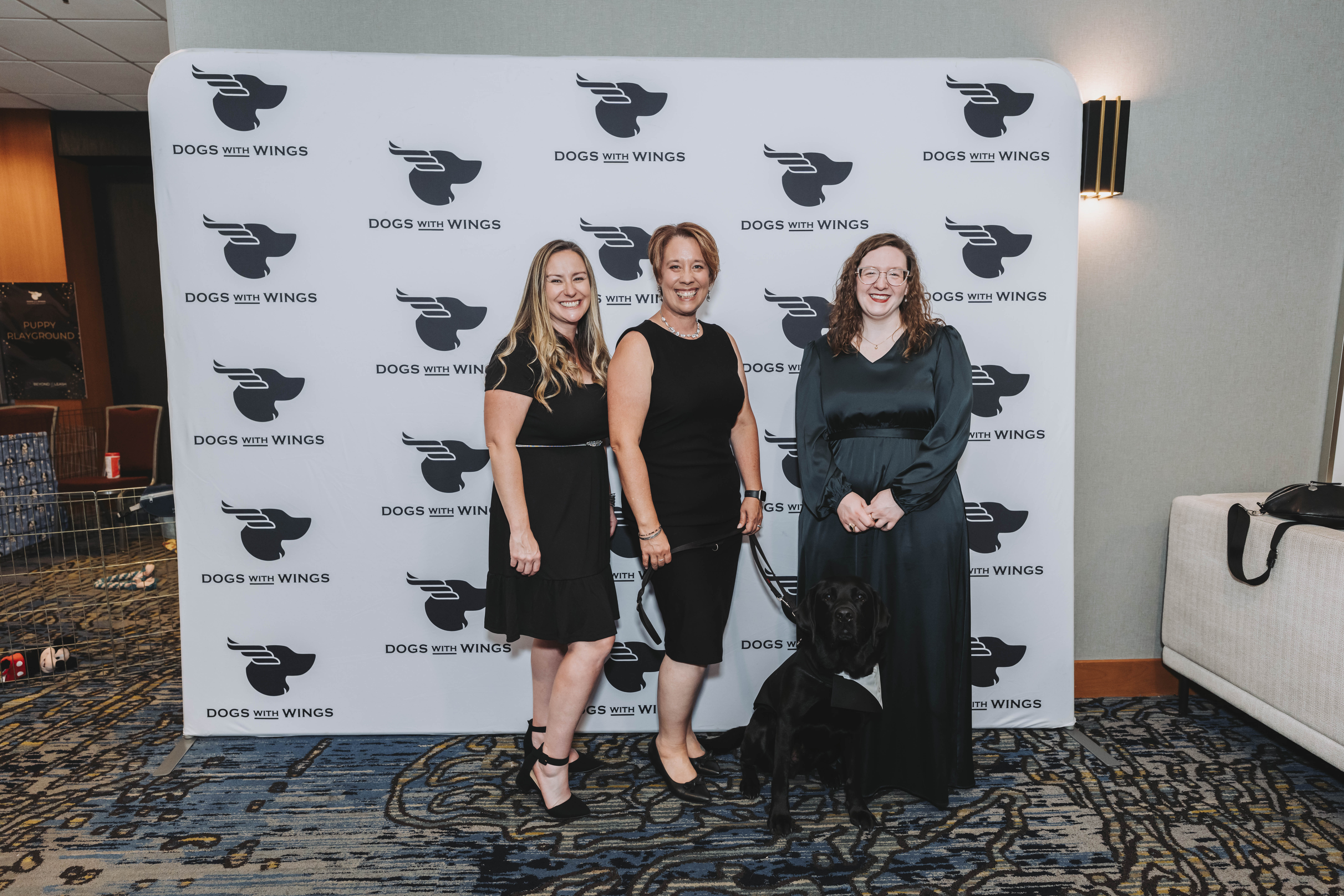 Three guests with a black Labrador in a tuxedo vest at the gala backdrop