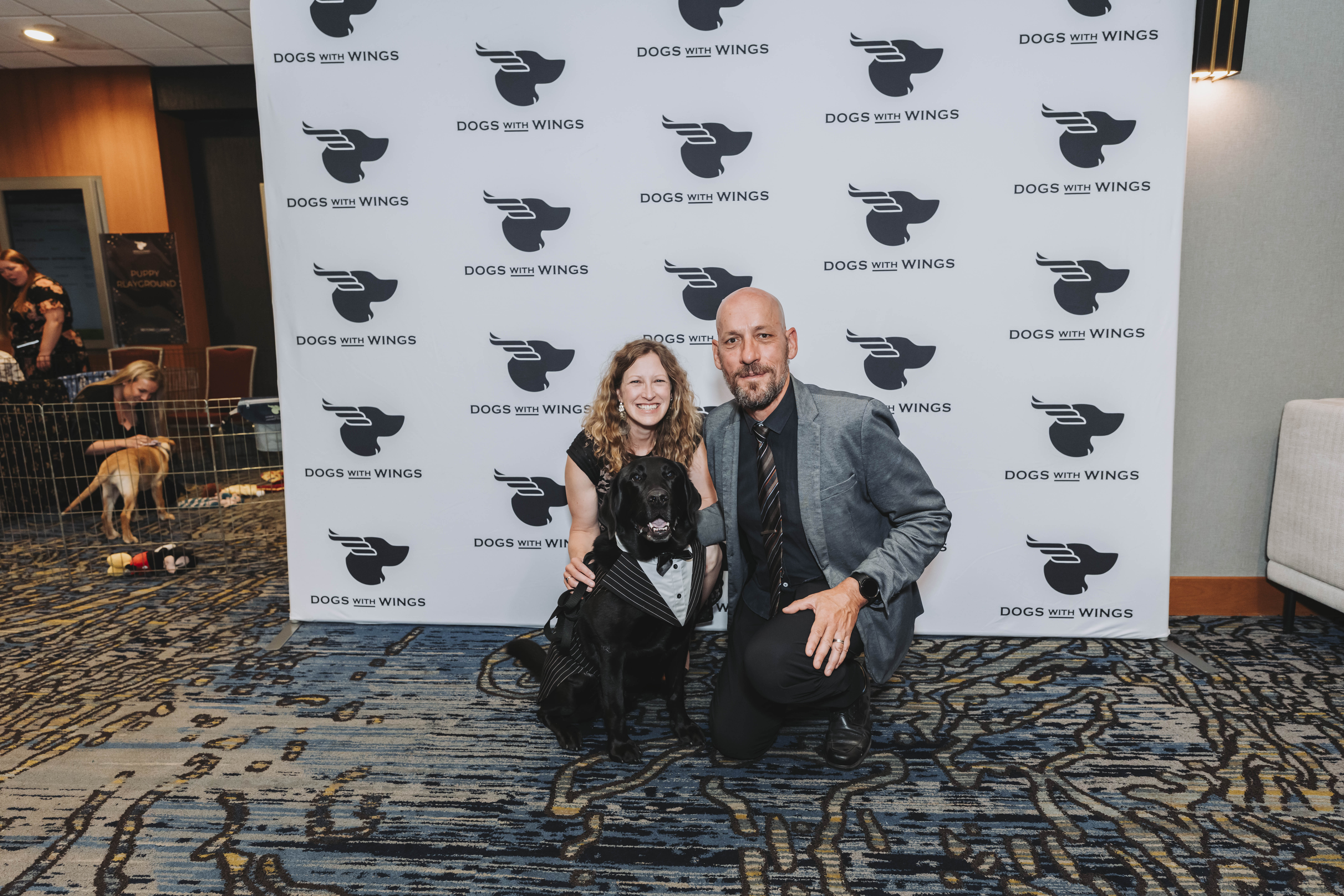 A couple kneeling with a black puppy in a tuxedo vest