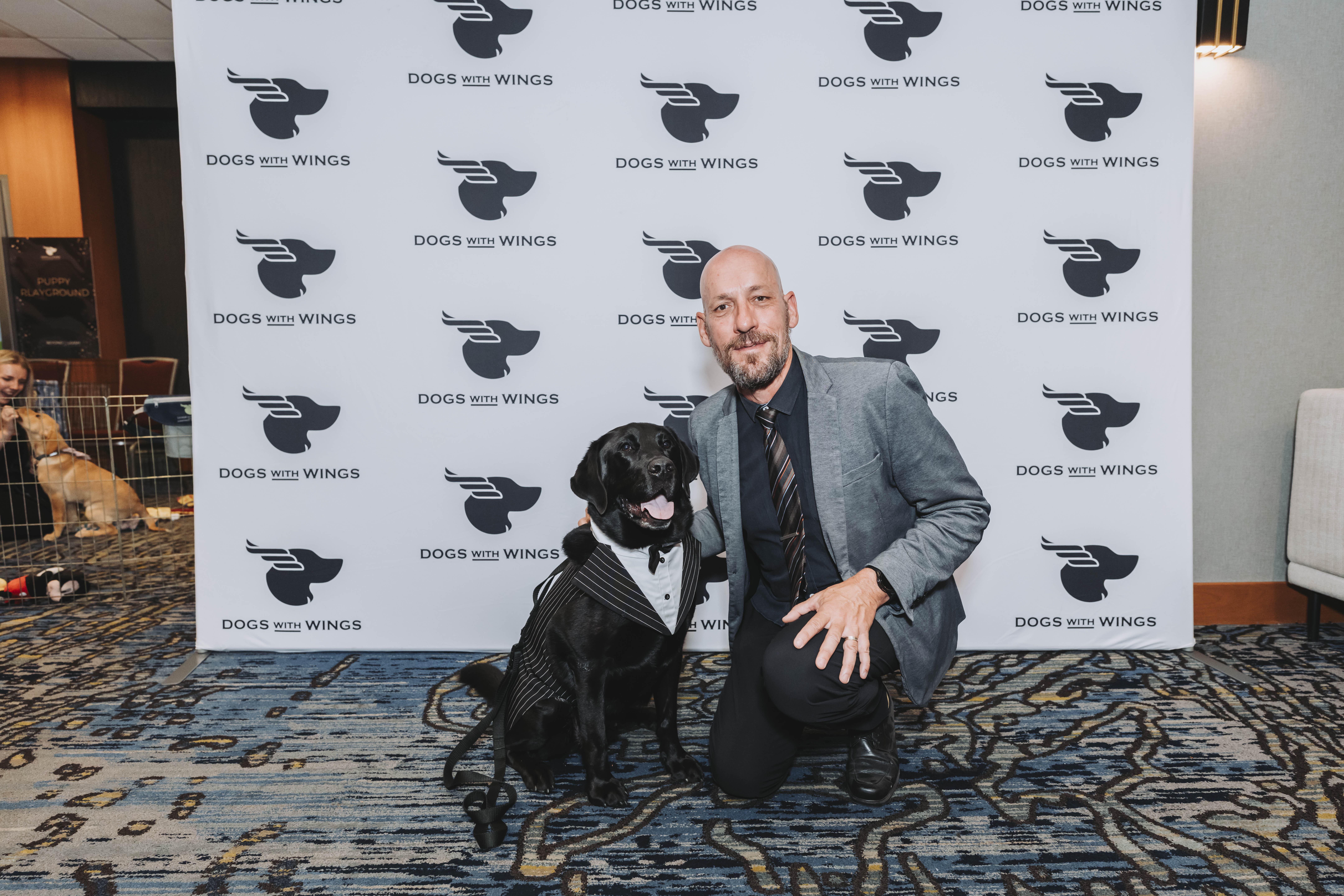 A guest kneeling with a black dog in a tuxedo harness at the backdrop