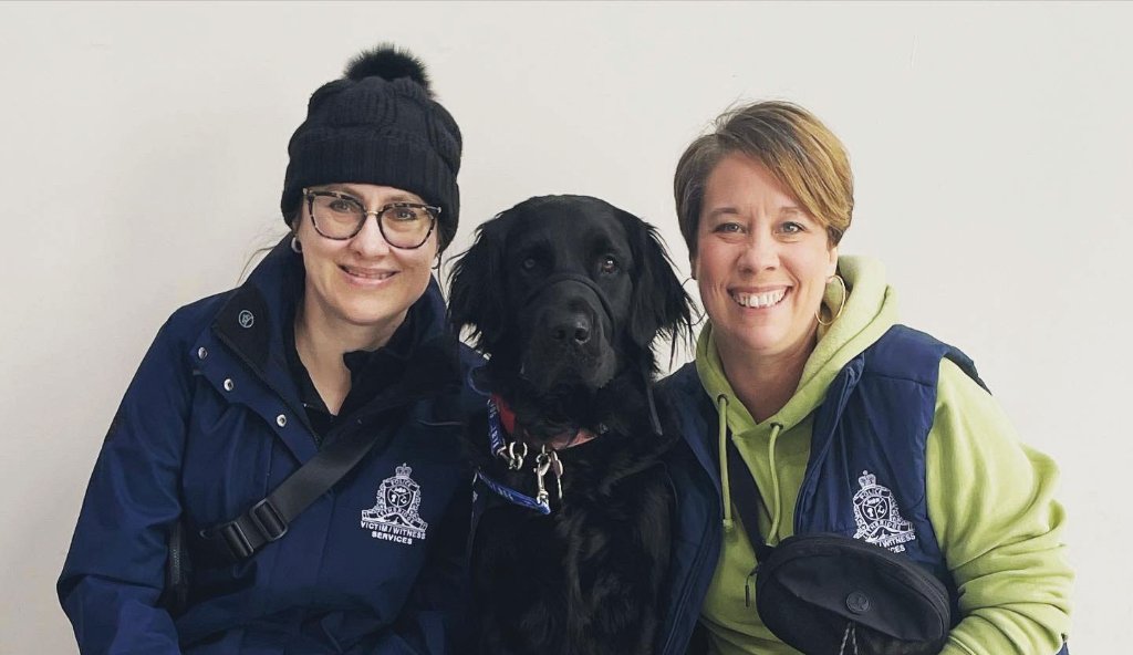 A black dog wearing a blue collar sits between two smiling women in professional attire, representing a facility service dog team