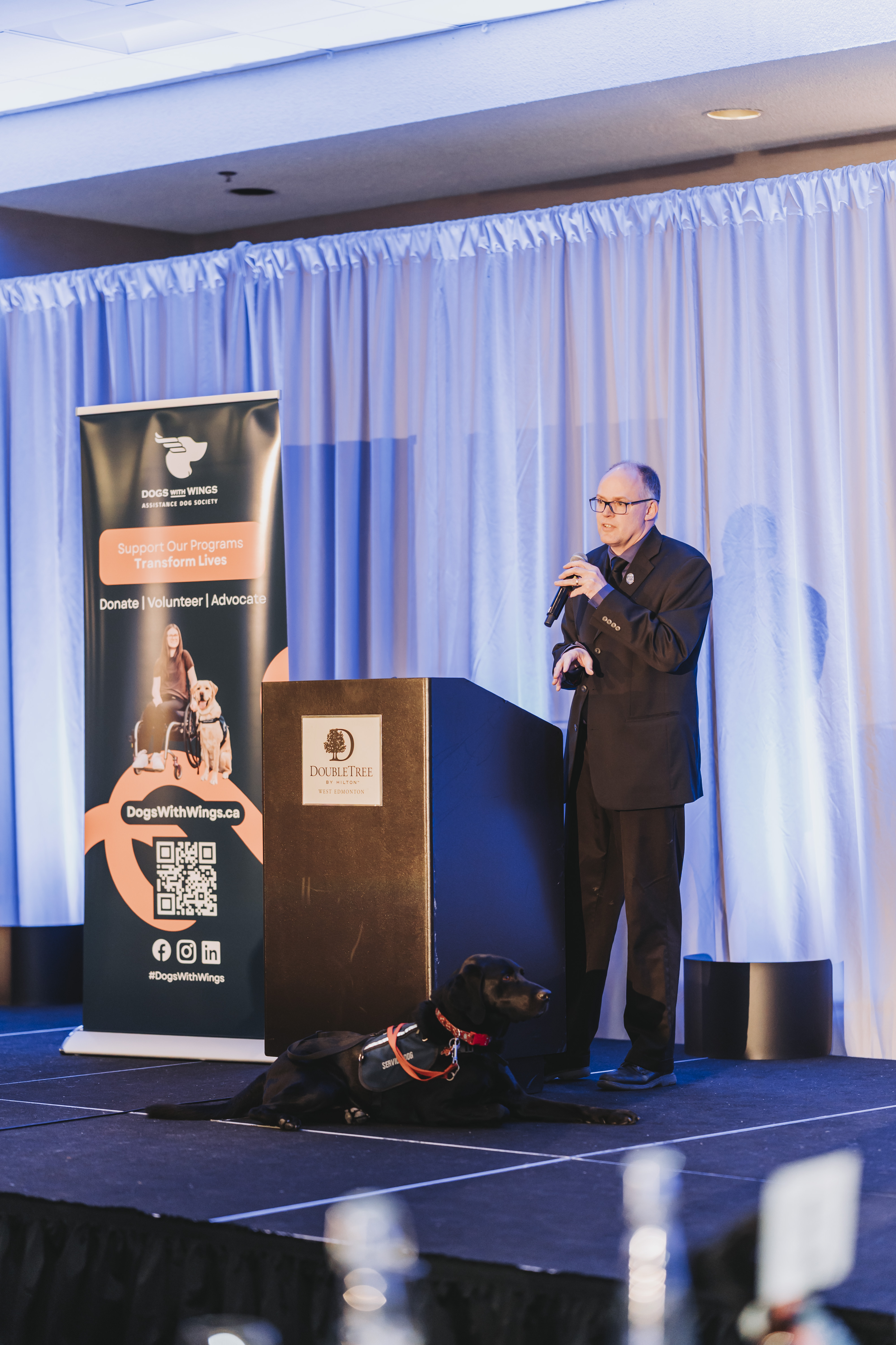 A guest speaking on stage with a black service dog resting nearby