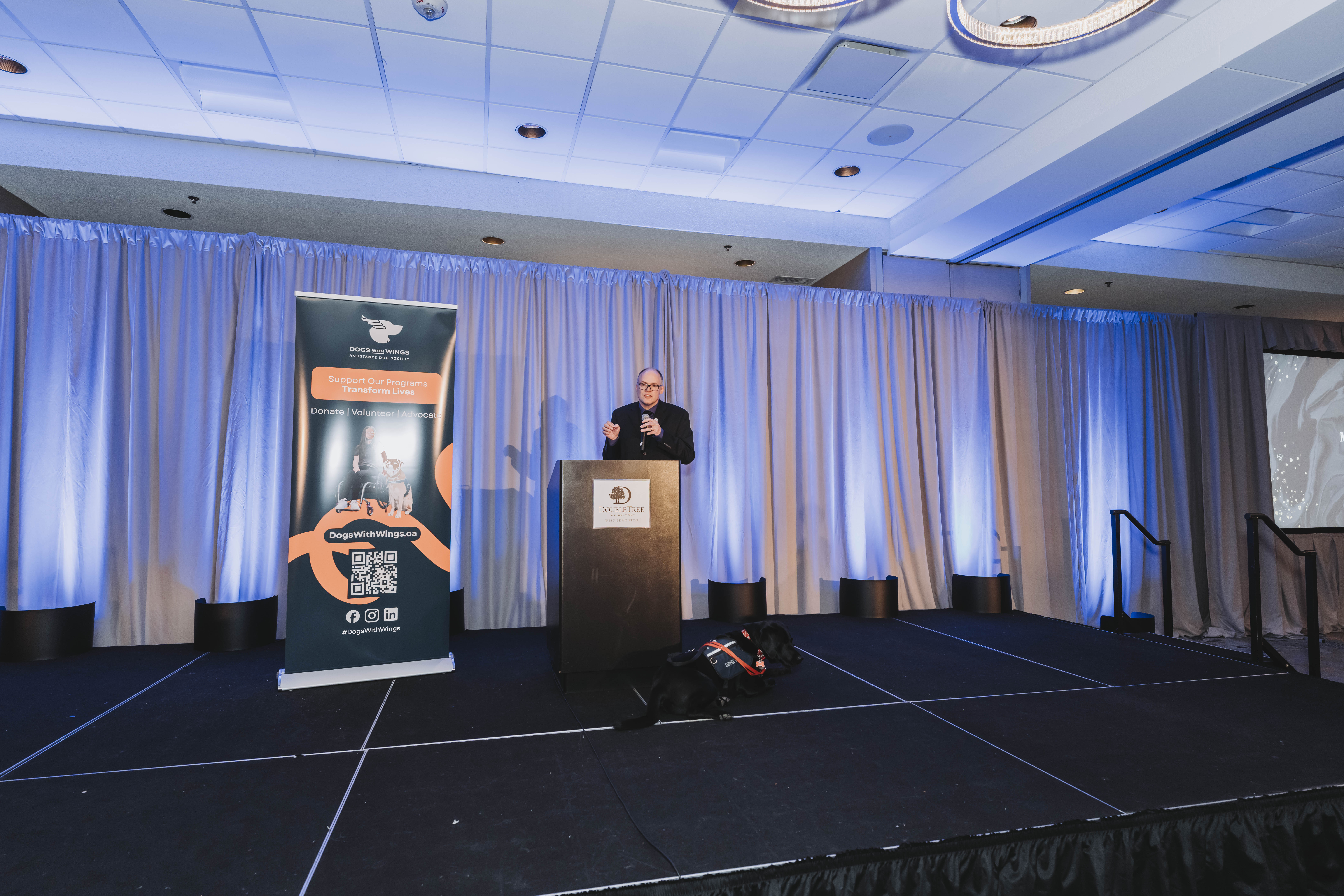 A guest speaking at the podium with a service dog on stage