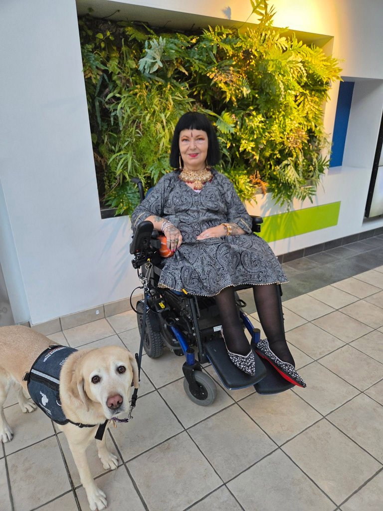 DWW Pixel, a yellow Labrador service dog wearing a vest, stands beside their partner who uses a power wheelchair, in front of a lush living wall
