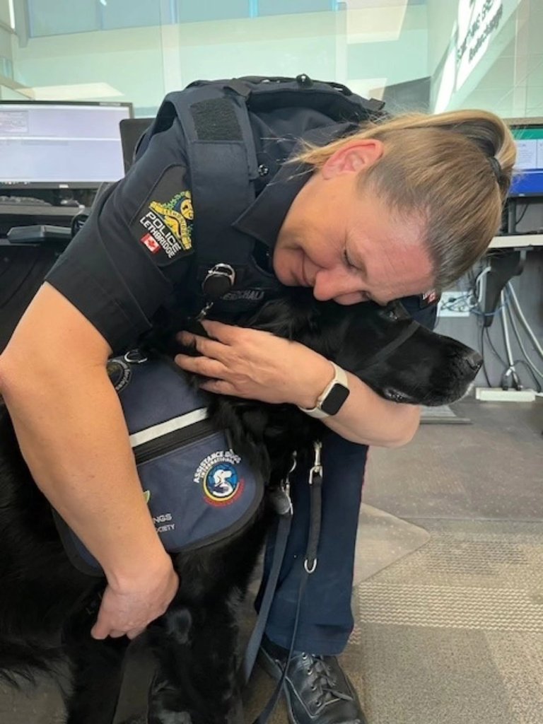 A Lethbridge Police Service officer embracing DWW Kourt, a black Labrador Accredited Facility Dog wearing a Dogs With Wings vest