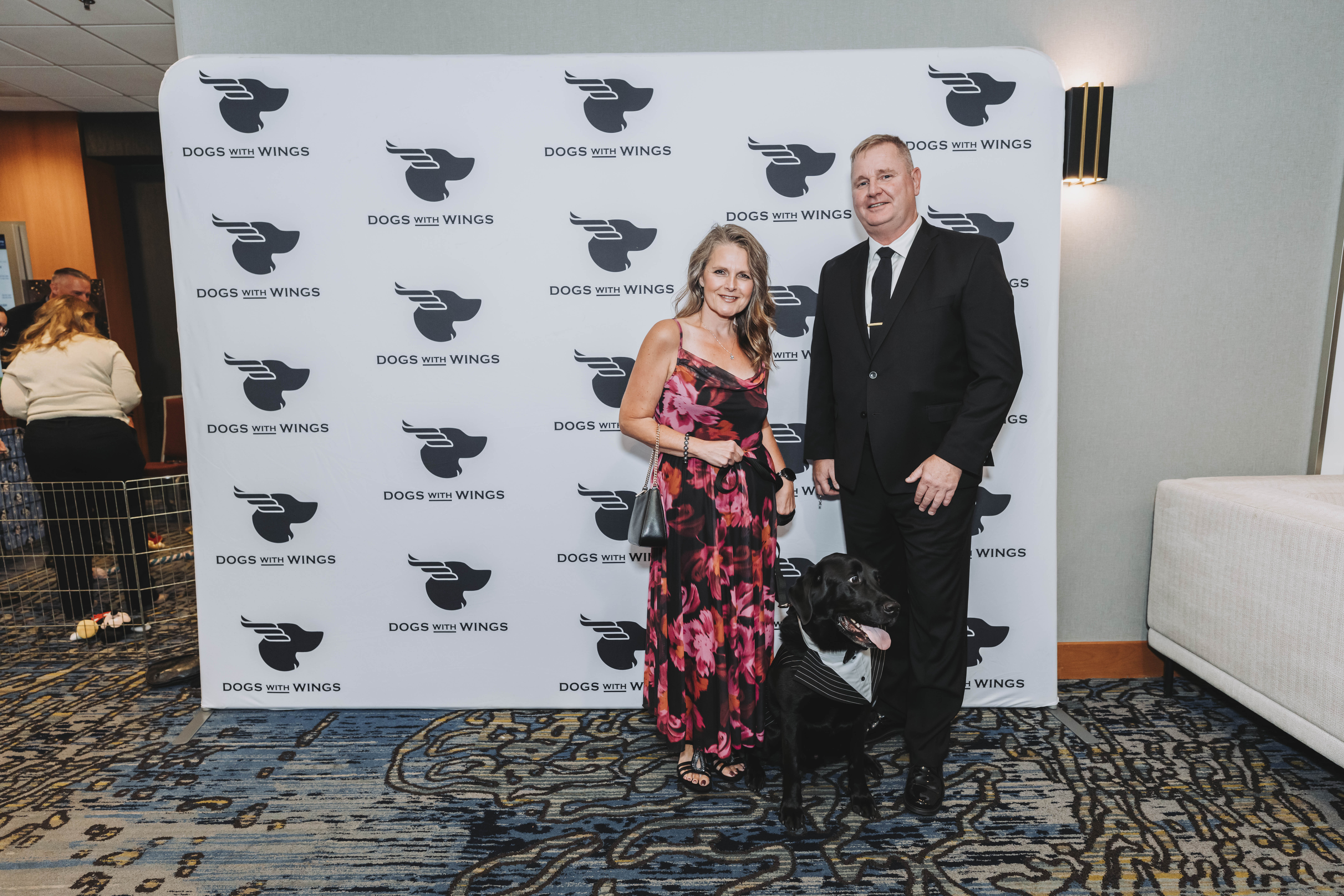 A couple posing with a black Labrador in a tuxedo harness
