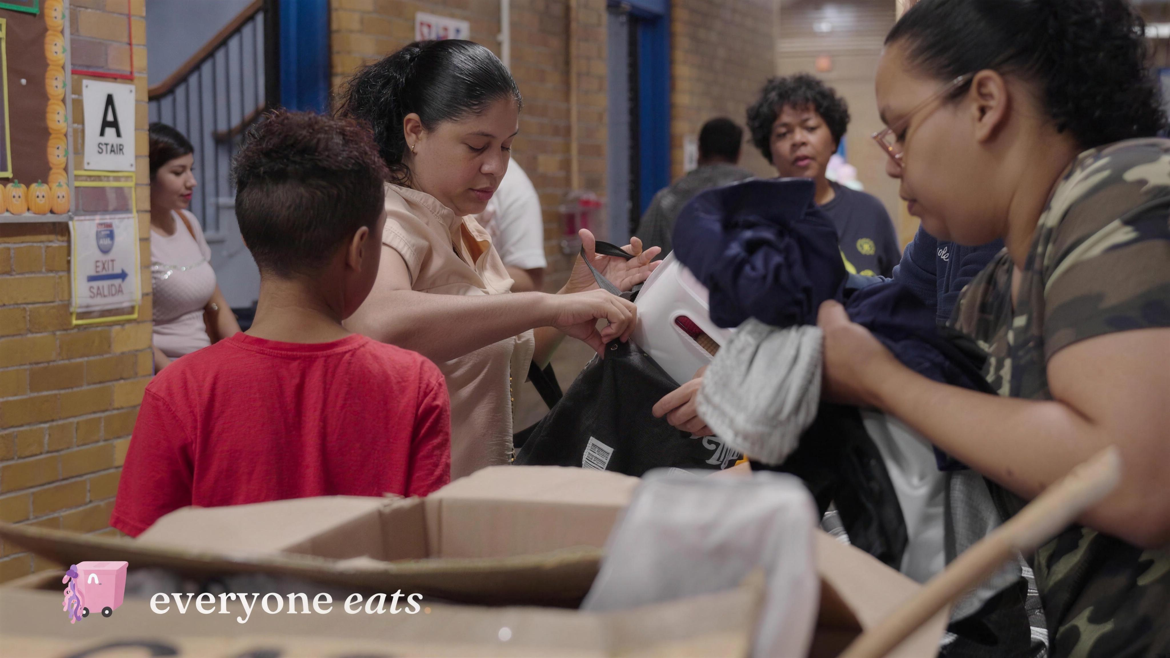 A volunteer handing folded essentials to a parent while a child looks on.