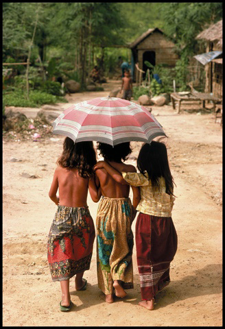 Three Cambodian children sharing an umbrella on a dirt path at Aranyaprathet refugee camp, Thailand
