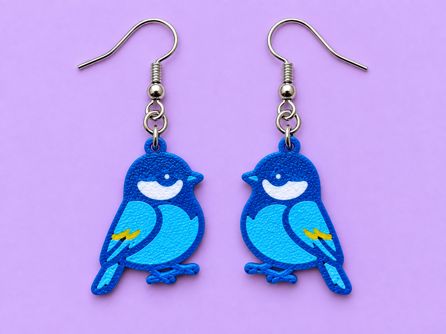 Blue tit bird earrings in royal blue with light blue wing patches and yellow accents, facing inward toward each other, on a light purple background