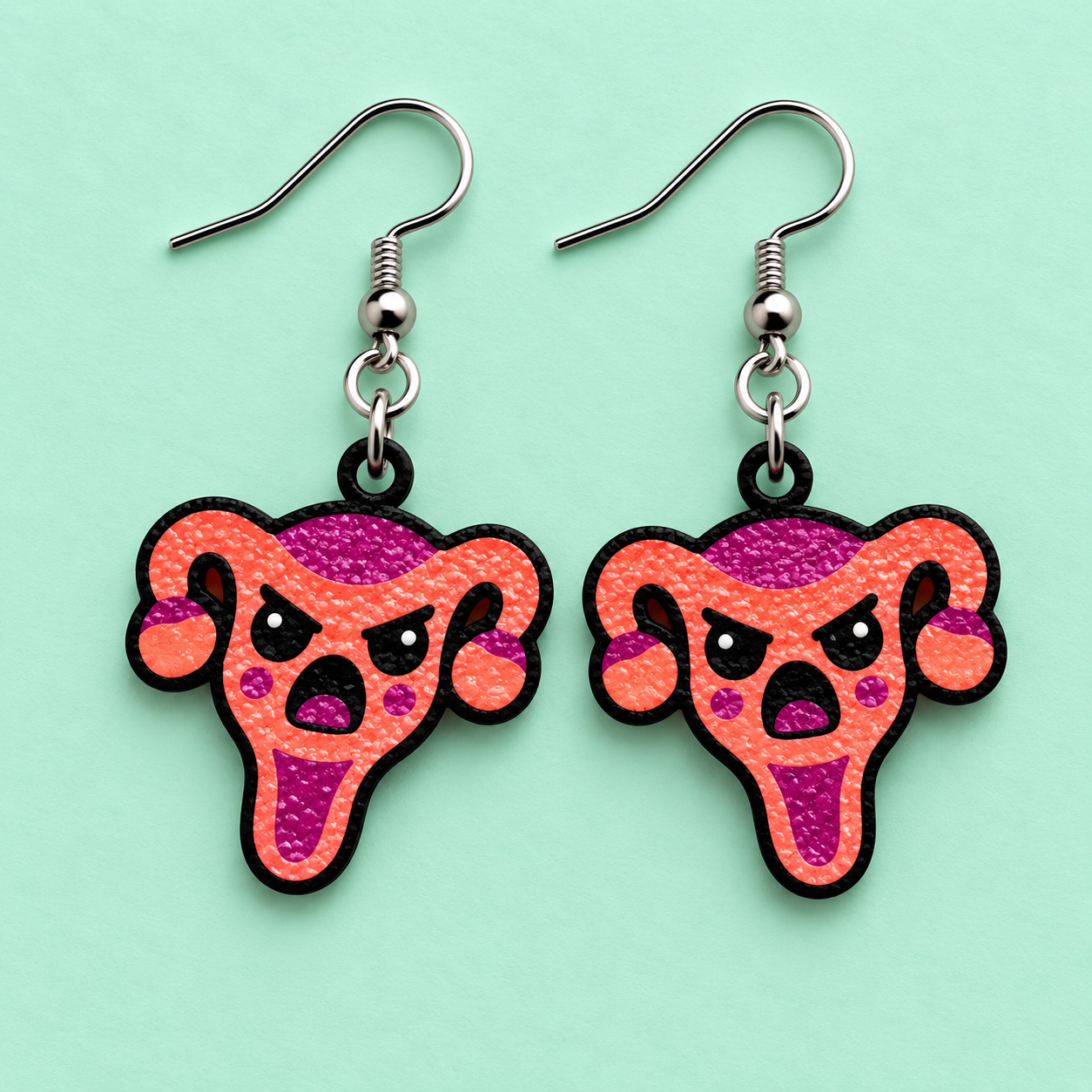 Angry uterus earrings in coral orange and magenta with black angry brows on a light mint green background