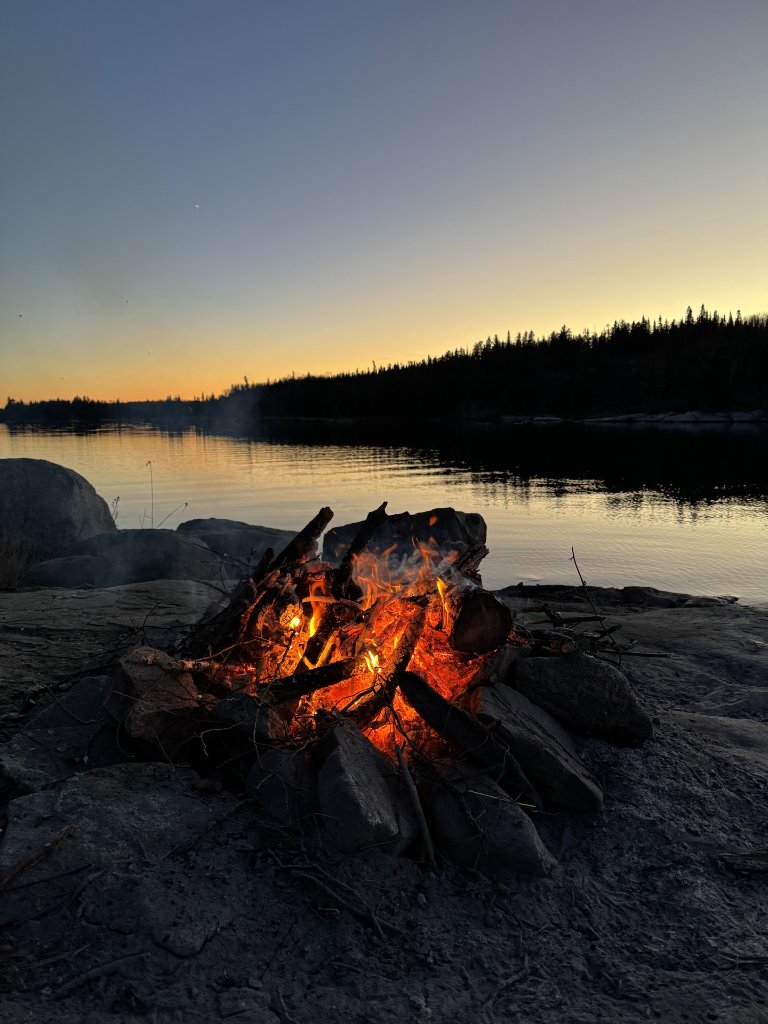 A crackling campfire on the rocks at the water's edge as the sky darkens