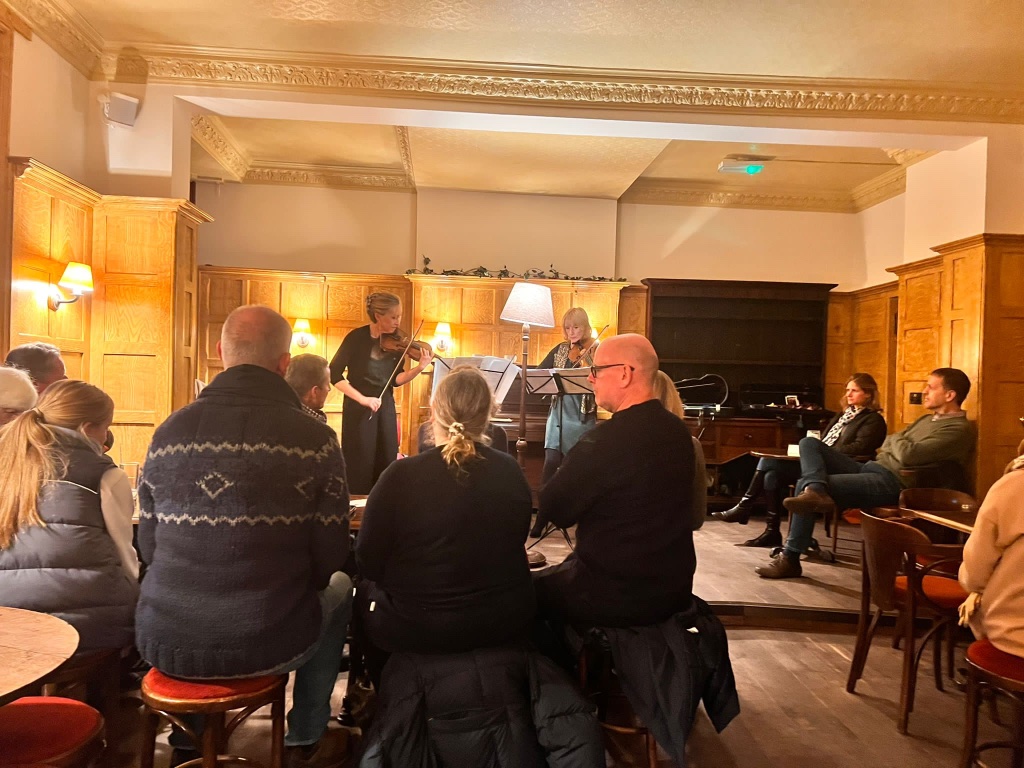 Two string players performing to a seated audience in the wood-panelled Flag Room