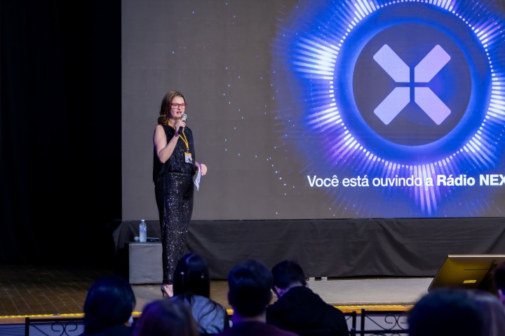 Palestrante no palco com tela da Rádio NEXO ao fundo
