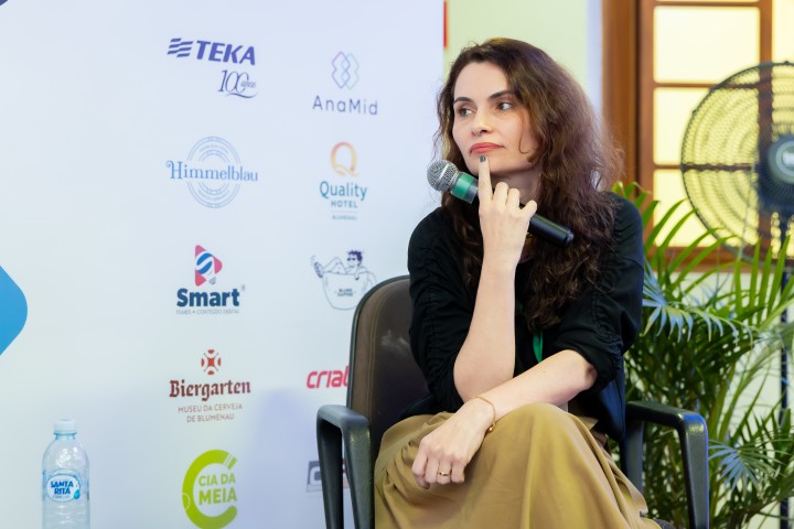 Palestrante sentada com mural de patrocinadores ao fundo