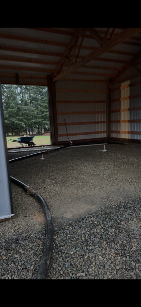 Interior of an unfinished pole barn with a leveled crushed-gravel base and utility stubs prepared for a concrete floor pour