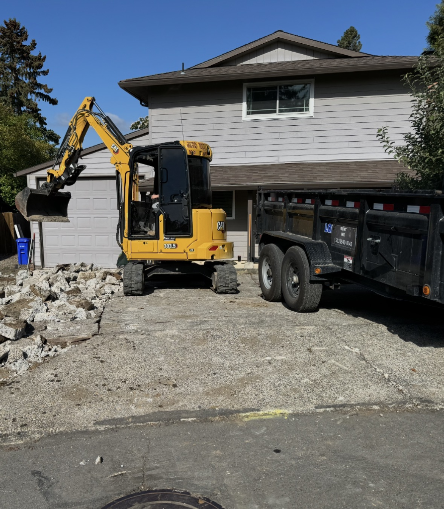 CAT mini-excavator demolishing an old concrete driveway with a dump trailer hitched nearby