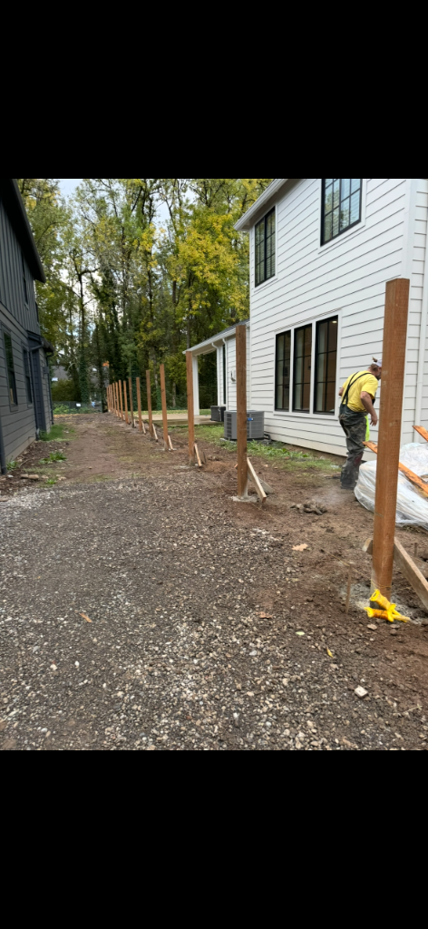 Vertical wooden fence posts installed in a line on a gravel site with a worker setting the first post
