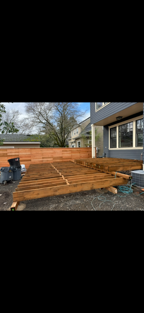 Elevated wooden deck joist framing ledgered to the side of a grey-sided house