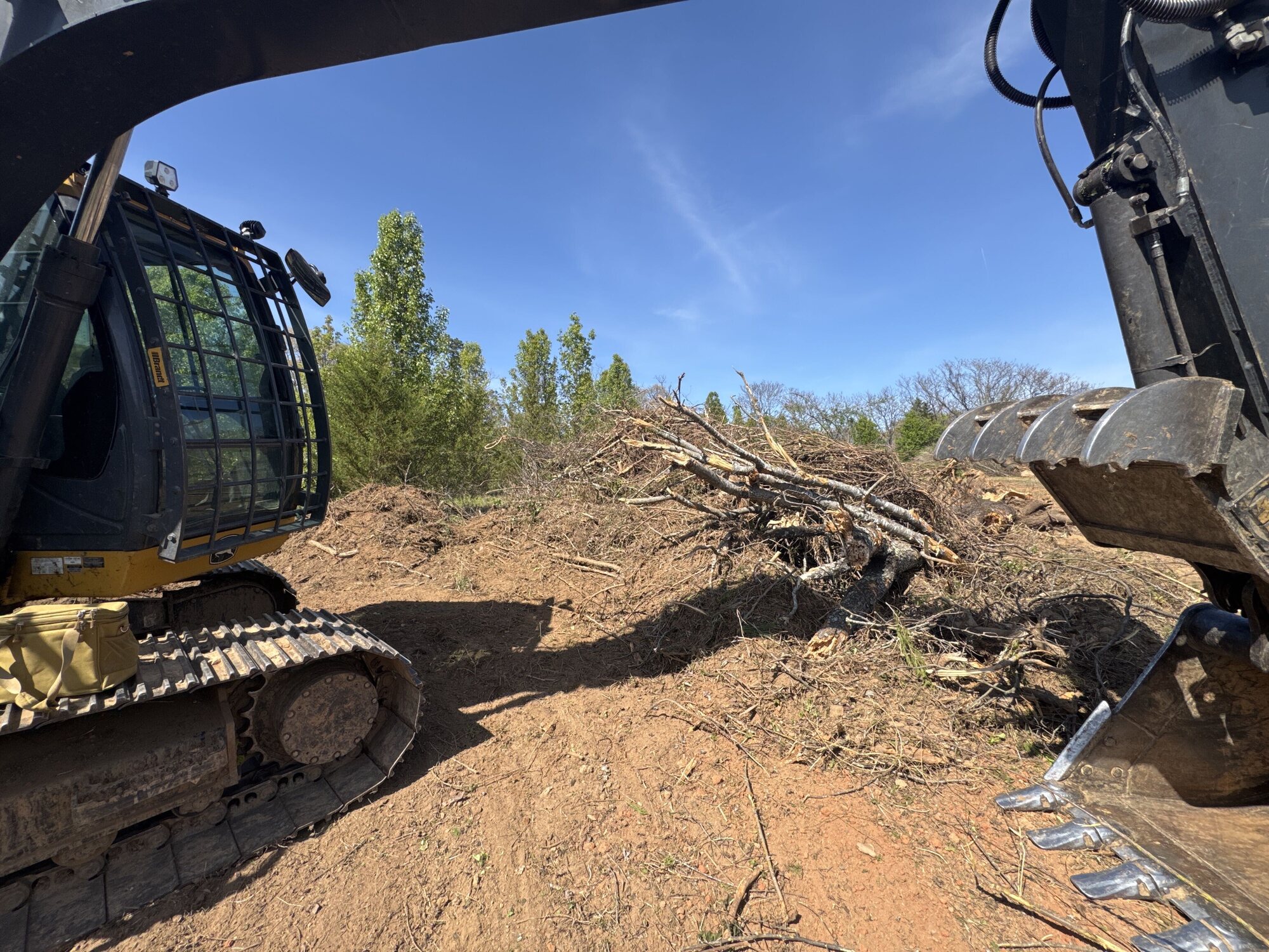 Excavator bucket and tracks beside a pile of cleared brush on a job site